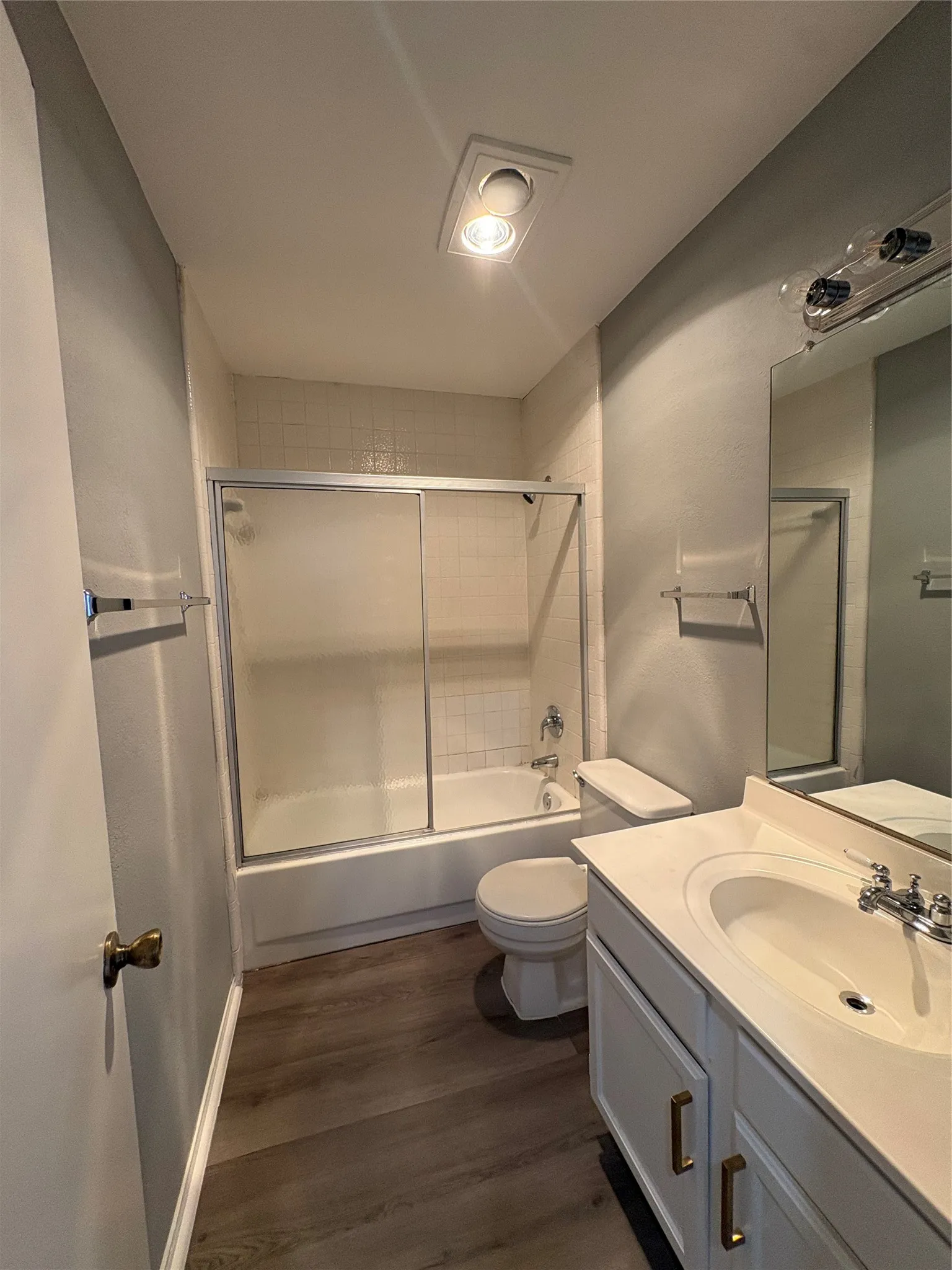 Bathroom featuring wood-finish flooring, white vanity with gold-tone hardware, integrated sink, and a shower-tub combination with sliding glass doors