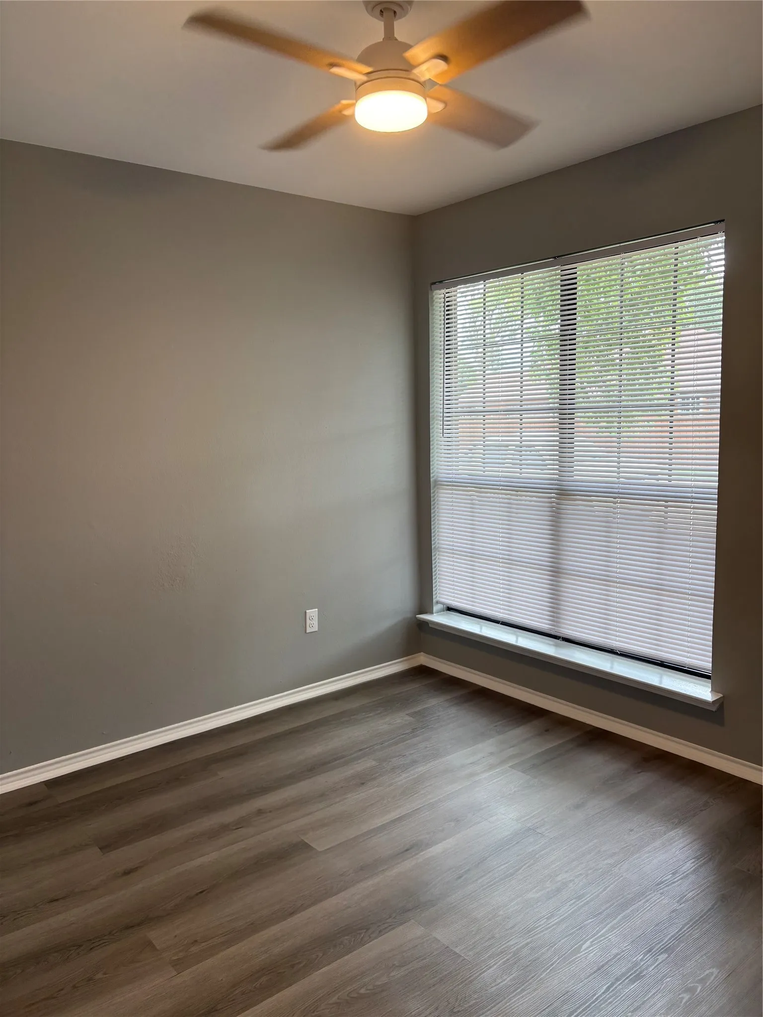 Wood-finish flooring, neutral wall tones, window with horizontal blinds, and a ceiling fan with integrated lighting
