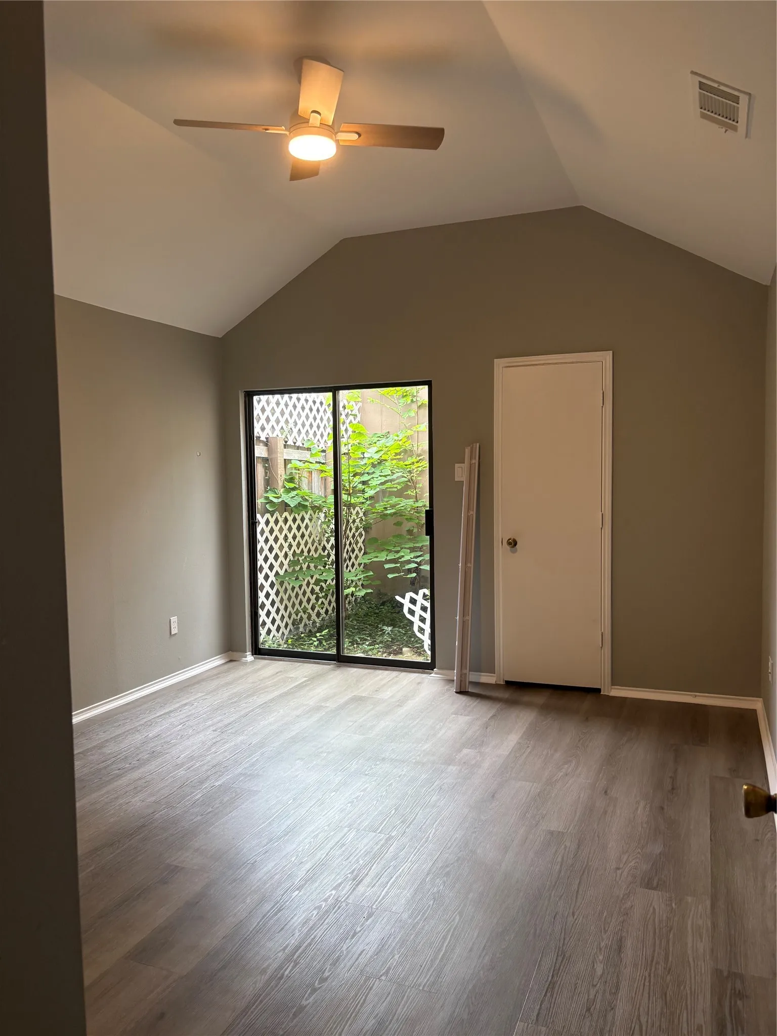 Vaulted ceiling, wood-finish flooring, ceiling fan with integrated lighting, sliding glass door, and a private patio with lattice fencing