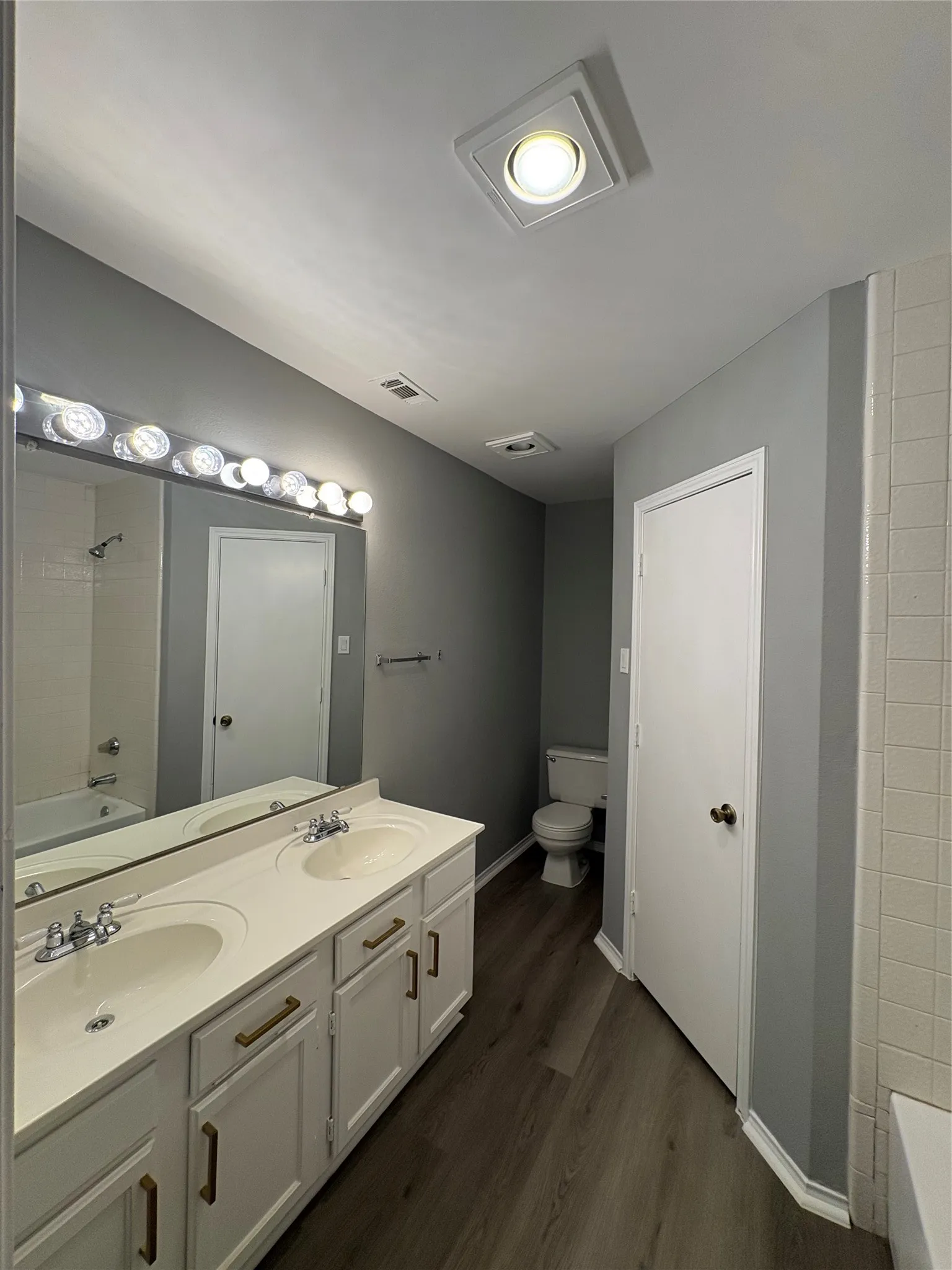 Dual vanity with integrated sinks, wood-finish flooring, and white cabinetry with brass-toned hardware