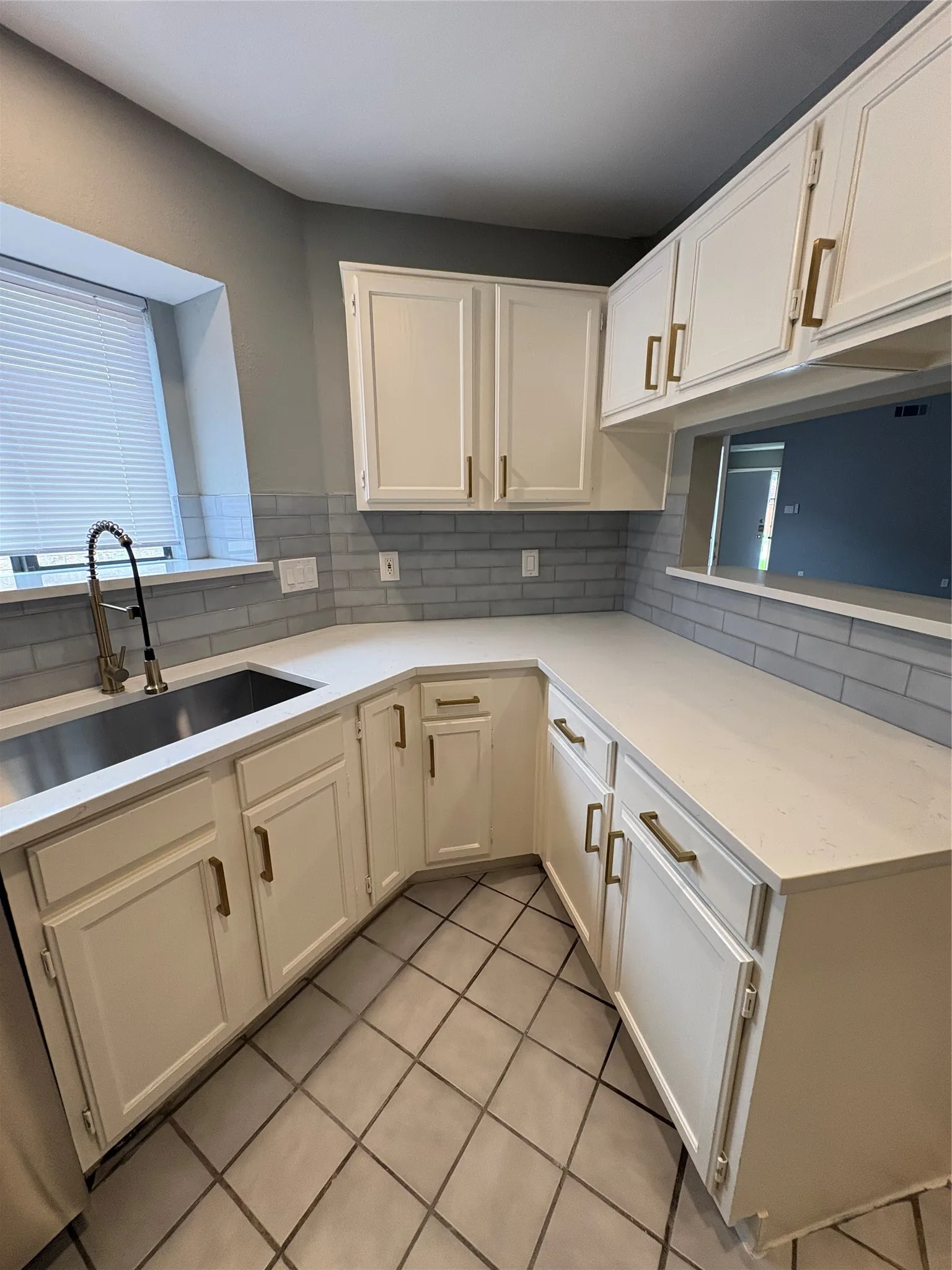 White cabinetry with gold-tone hardware, light-toned countertops, subway tile backsplash, stainless steel sink, and gray tiled flooring