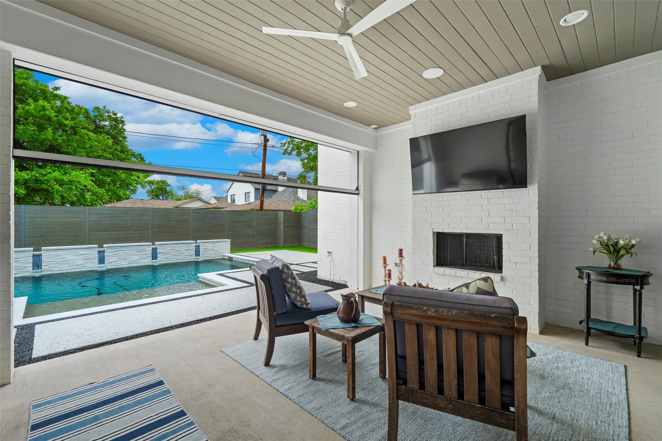 Cozy outdoor patio leading to the pool/spa area with electric roll-down screen, wooden ceiling, ceiling fan, and a brick fireplace.  Also features built-in grill!