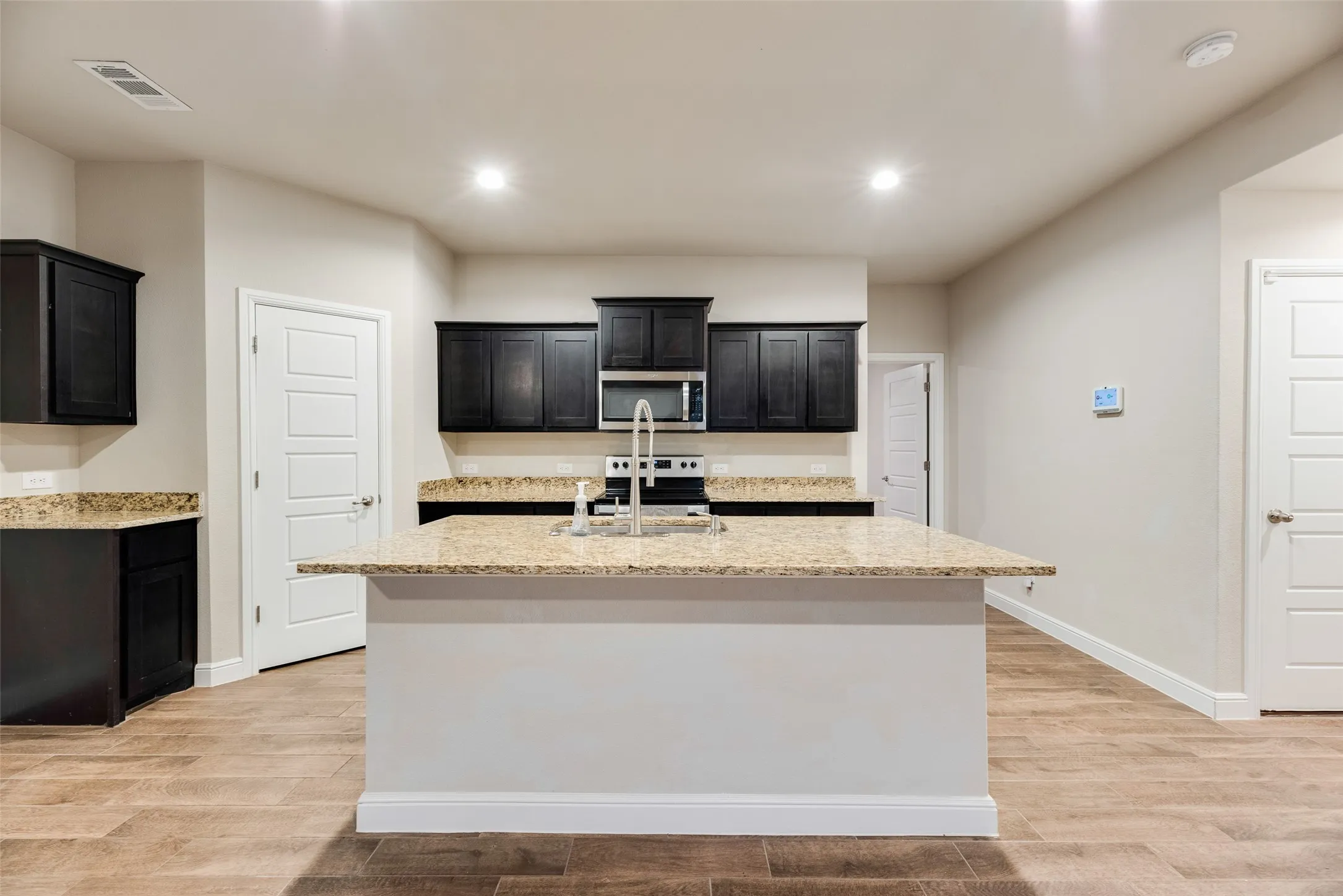 Kitchen featuring an island with a stainless steel sink, black cabinetry, speckled countertops, and wood-finish flooring