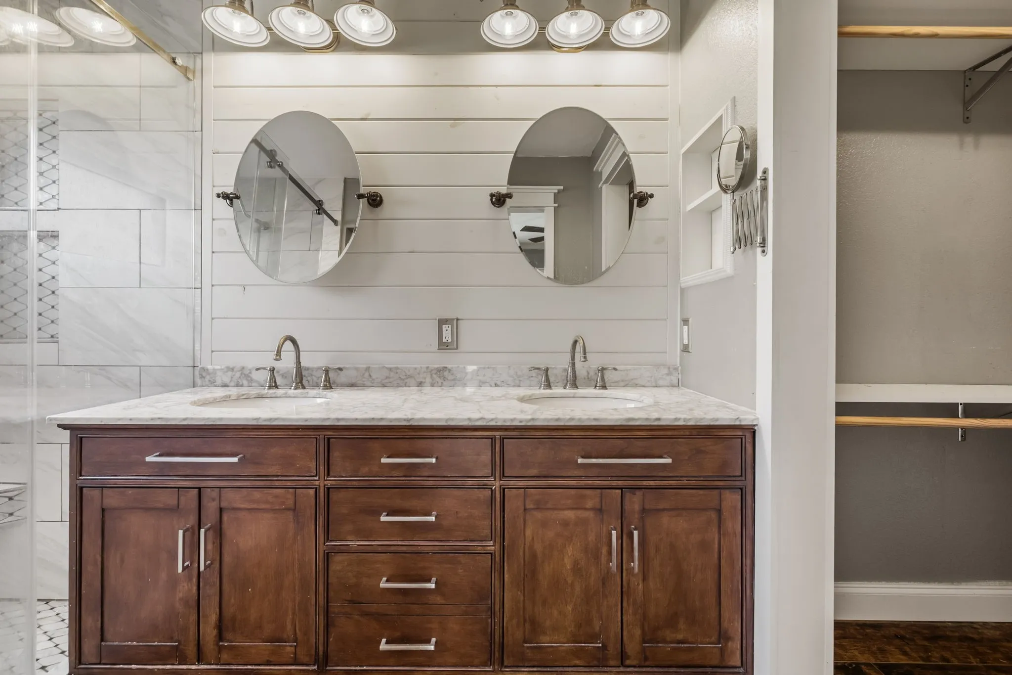 Dual sink vanity with dark wood-finish cabinetry, light-toned countertop, and individual oval mirrors. Large walk-in shower, and walk-in closet is adjacent.