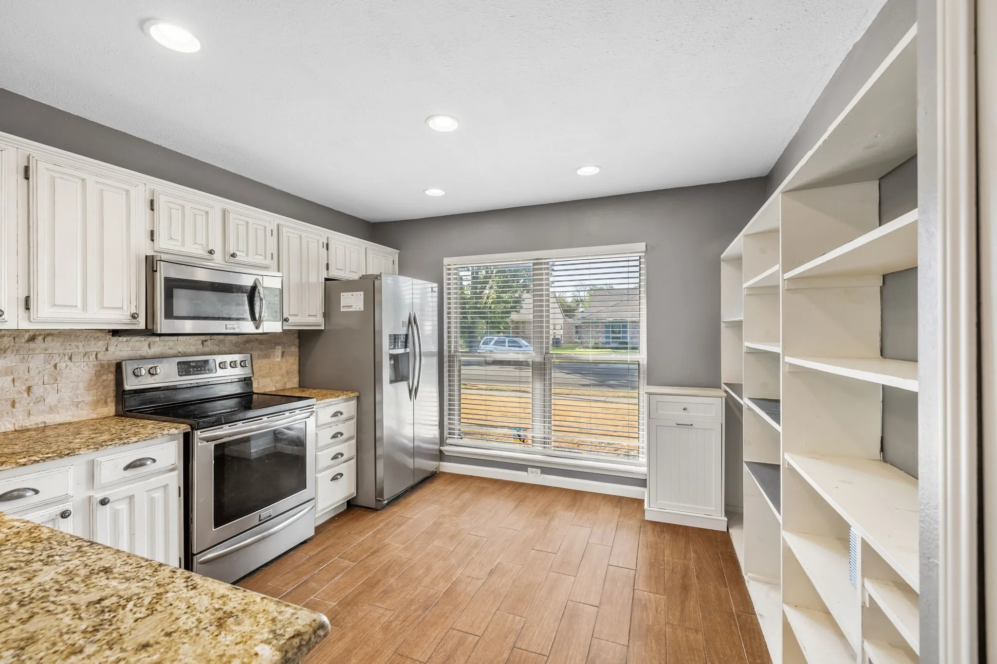 Kitchen featuring white cabinetry, granite countertops, stainless steel appliances, built-in microwave, pantry, abundance of open shelving, and LVP flooring. Large picture window allows natural light to fill the space.