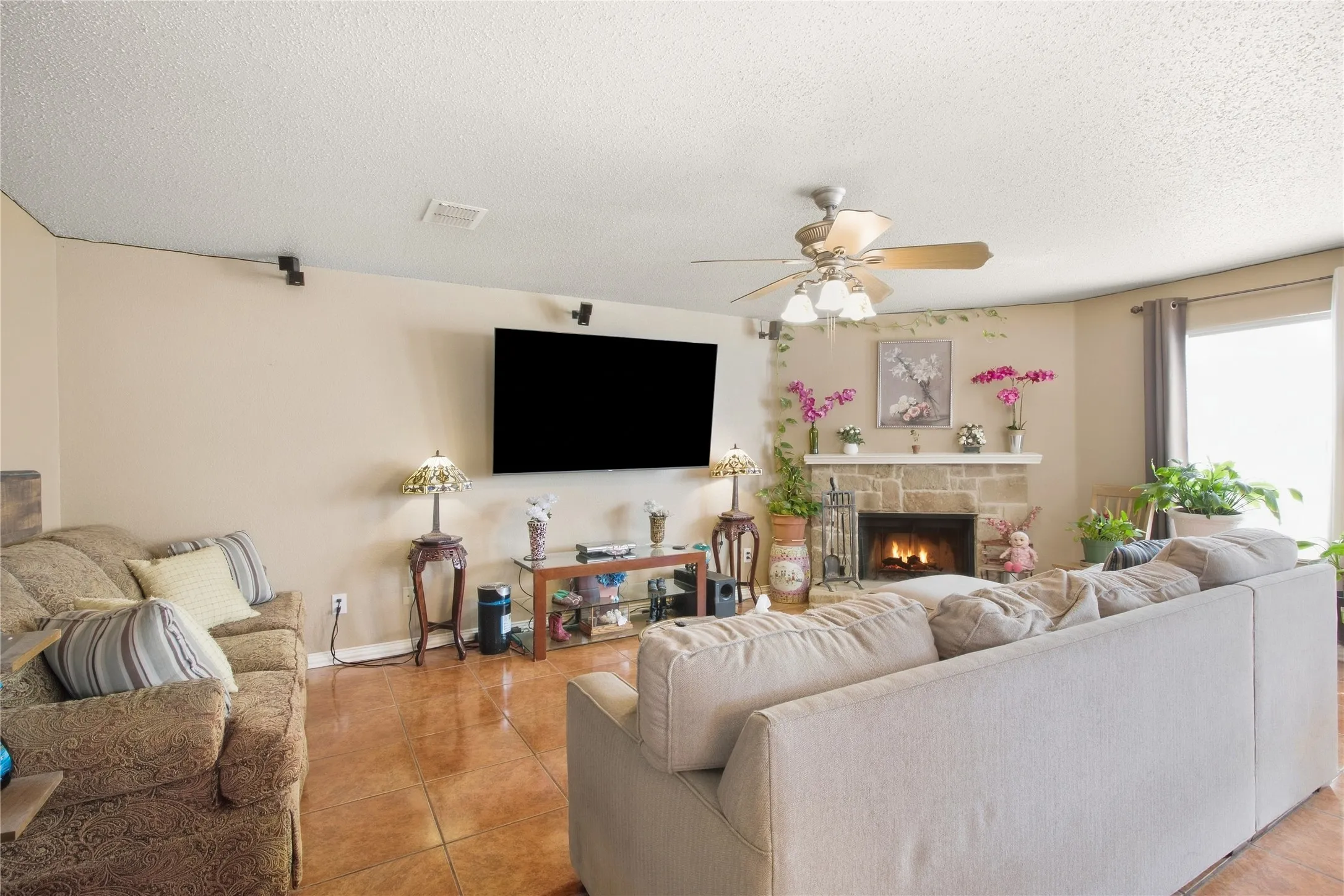 Tiled flooring, ceiling fan with integrated lighting, stone-faced fireplace, large window, and track lighting