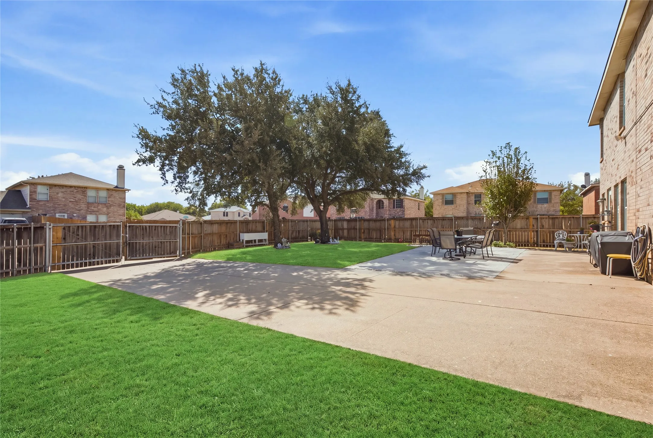 Expansive backyard featuring a large concrete patio, manicured lawn, and mature trees, enclosed by a wood fence with a double gate