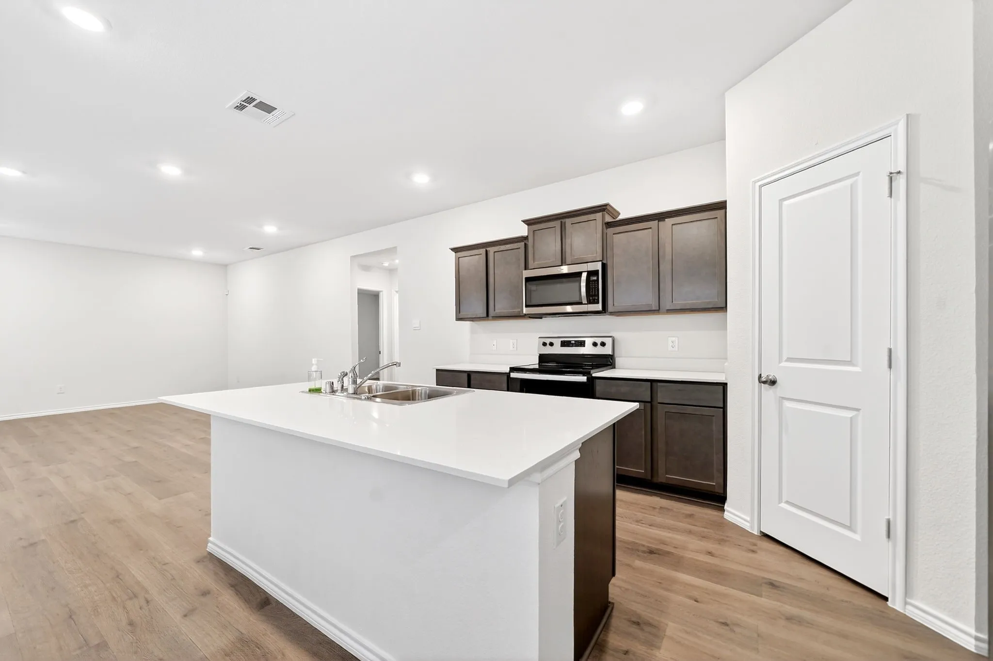 Kitchen island with an undermount sink and white countertop, dark wood-finish cabinetry, stainless steel microwave, and wood-finish flooring