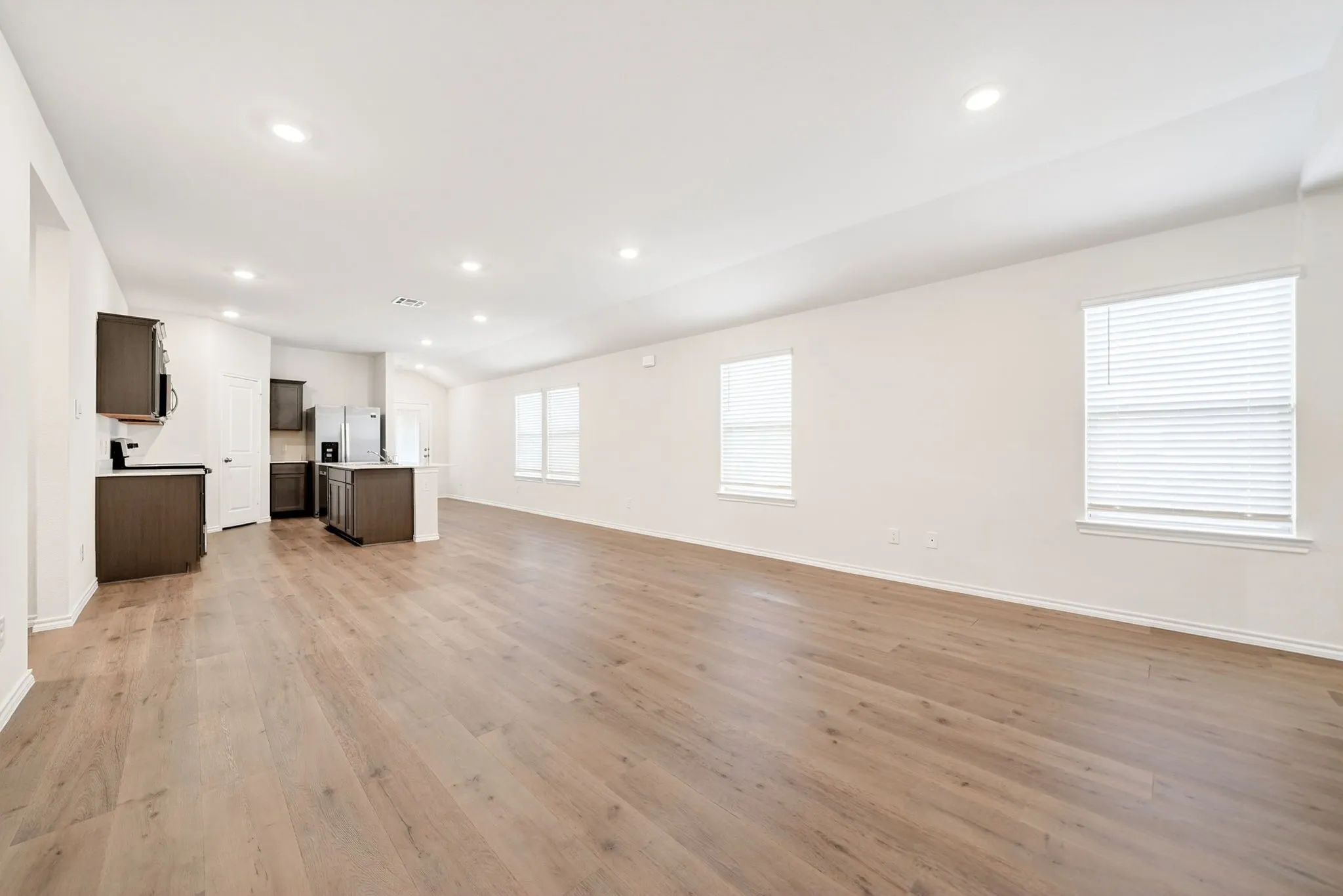 Expansive open-concept living area with wood-finish flooring, recessed lighting, multiple windows with blinds, and a kitchen featuring dark wood cabinetry and stainless steel appliances