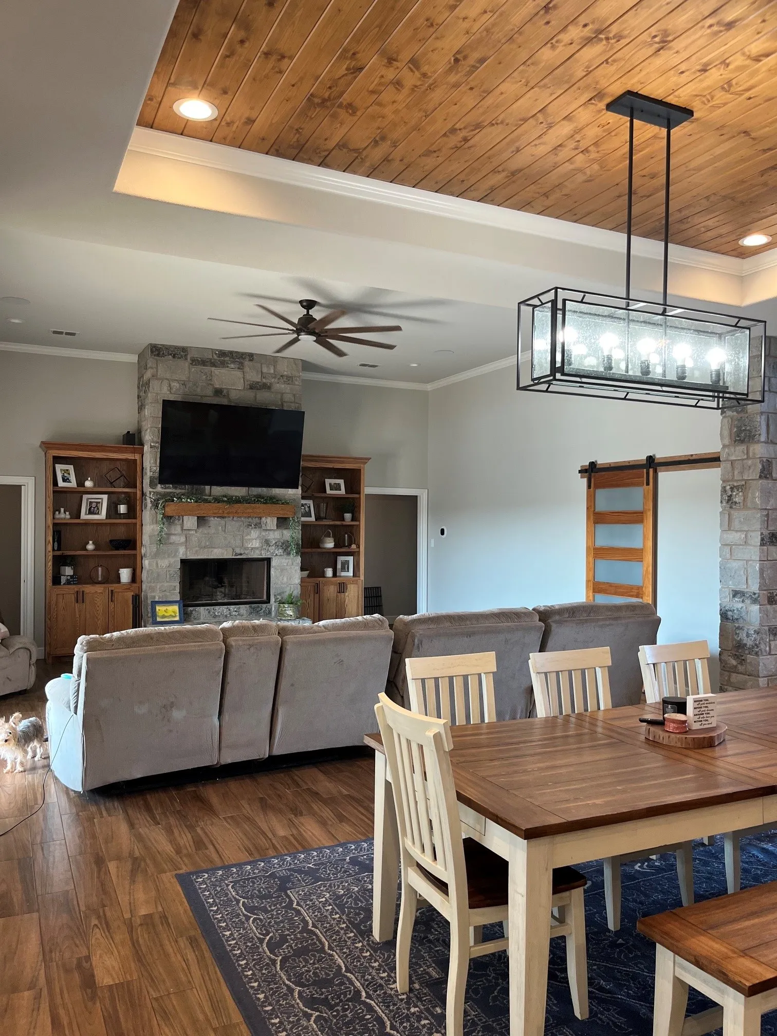 Wood-finish flooring throughout, tray ceiling with wood paneling, stone fireplace with a wood mantle, custom built-in shelving, and a wood barn door