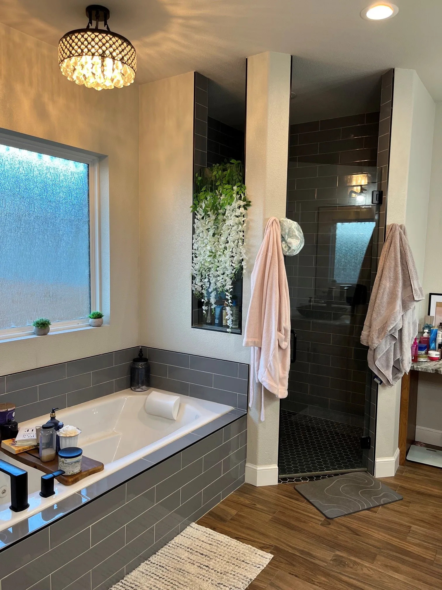 Soaking tub with subway tile surround, separate enclosed shower with dark tile, wood-finish flooring, and a decorative ceiling light fixture