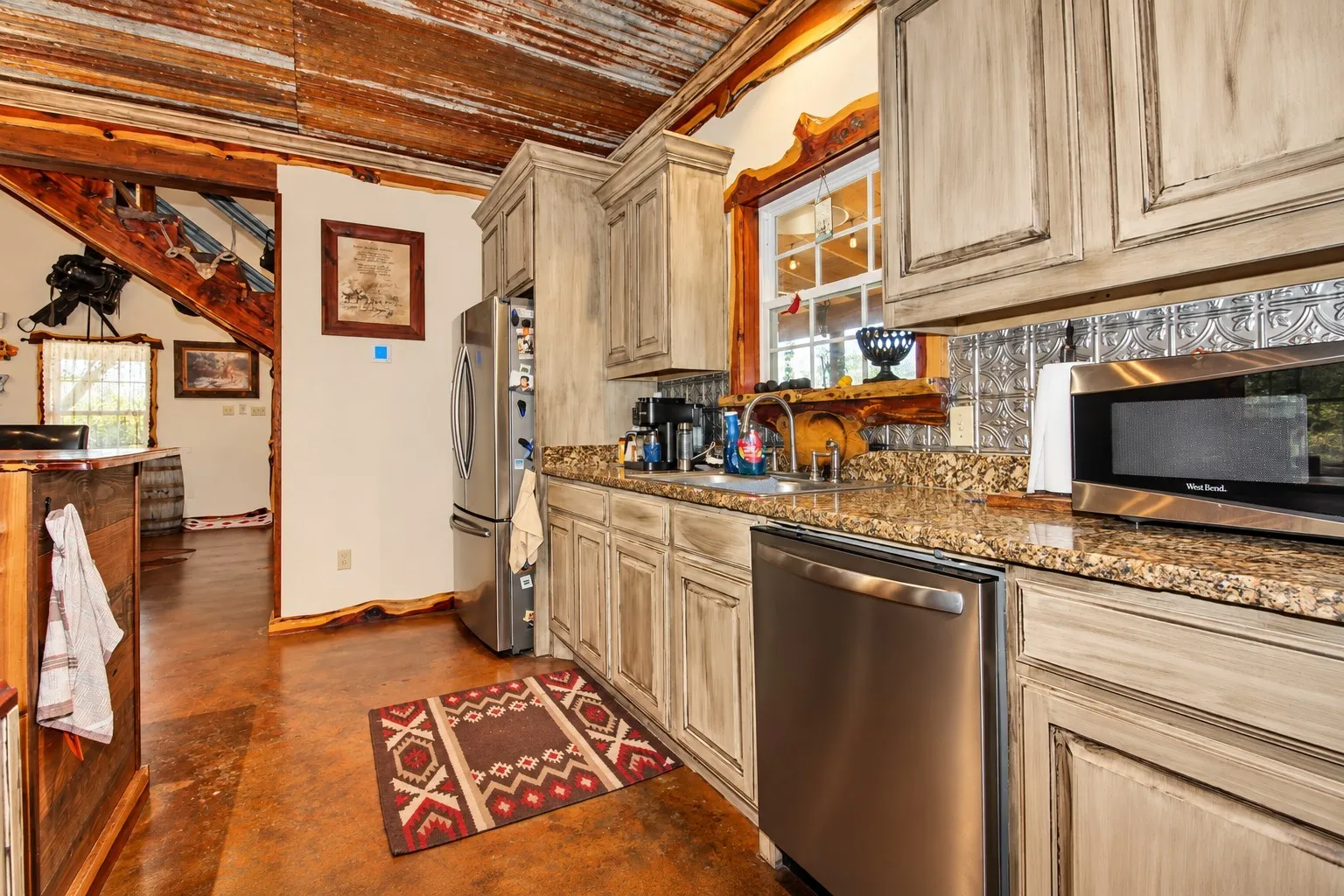 Kitchen featuring distressed cabinetry, granite countertops, stainless steel appliances, a decorative metal backsplash, and a corrugated metal ceiling