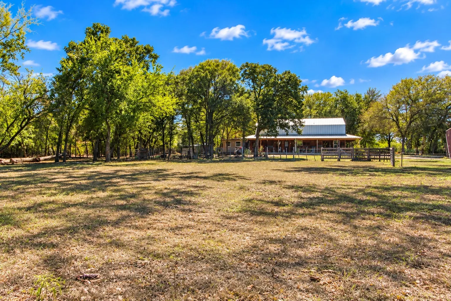 Expansive open field with scattered mature trees and a distant structure featuring a covered porch and metal roof