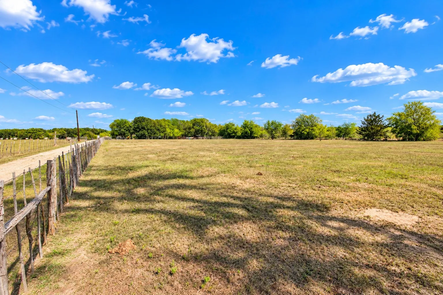 Expansive open field with scattered trees and a natural wood-post fence bordering a gravel path