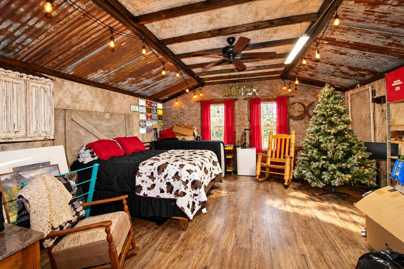 Vaulted ceiling with exposed wood beams and corrugated metal panels, wood-finish flooring, two windows with red drapes, and a ceiling fan with integrated lighting