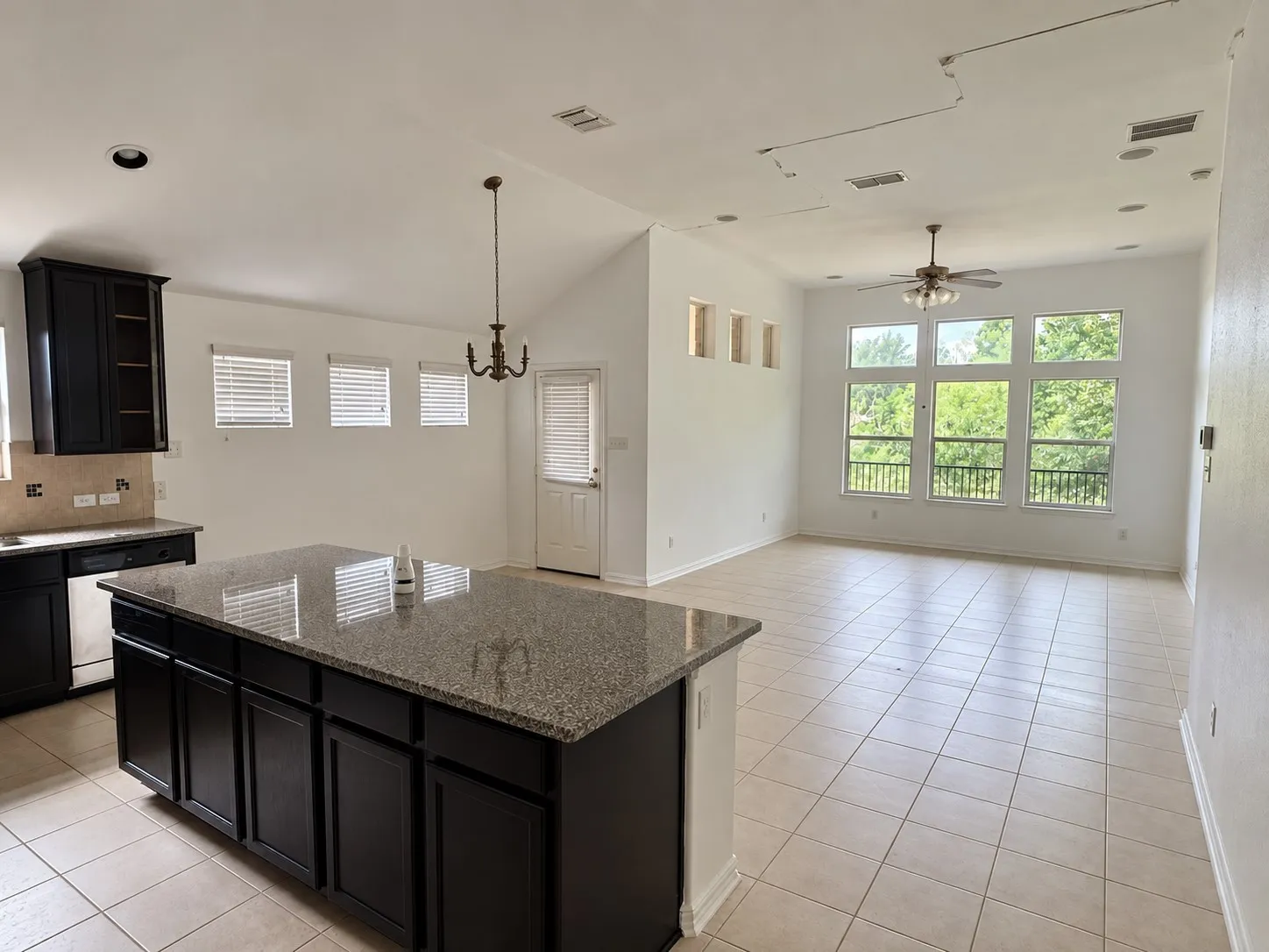 Open-concept living space with tiled flooring, dark wood-finish cabinetry, a large kitchen island with a stone countertop, and a wall of windows featuring decorative railing