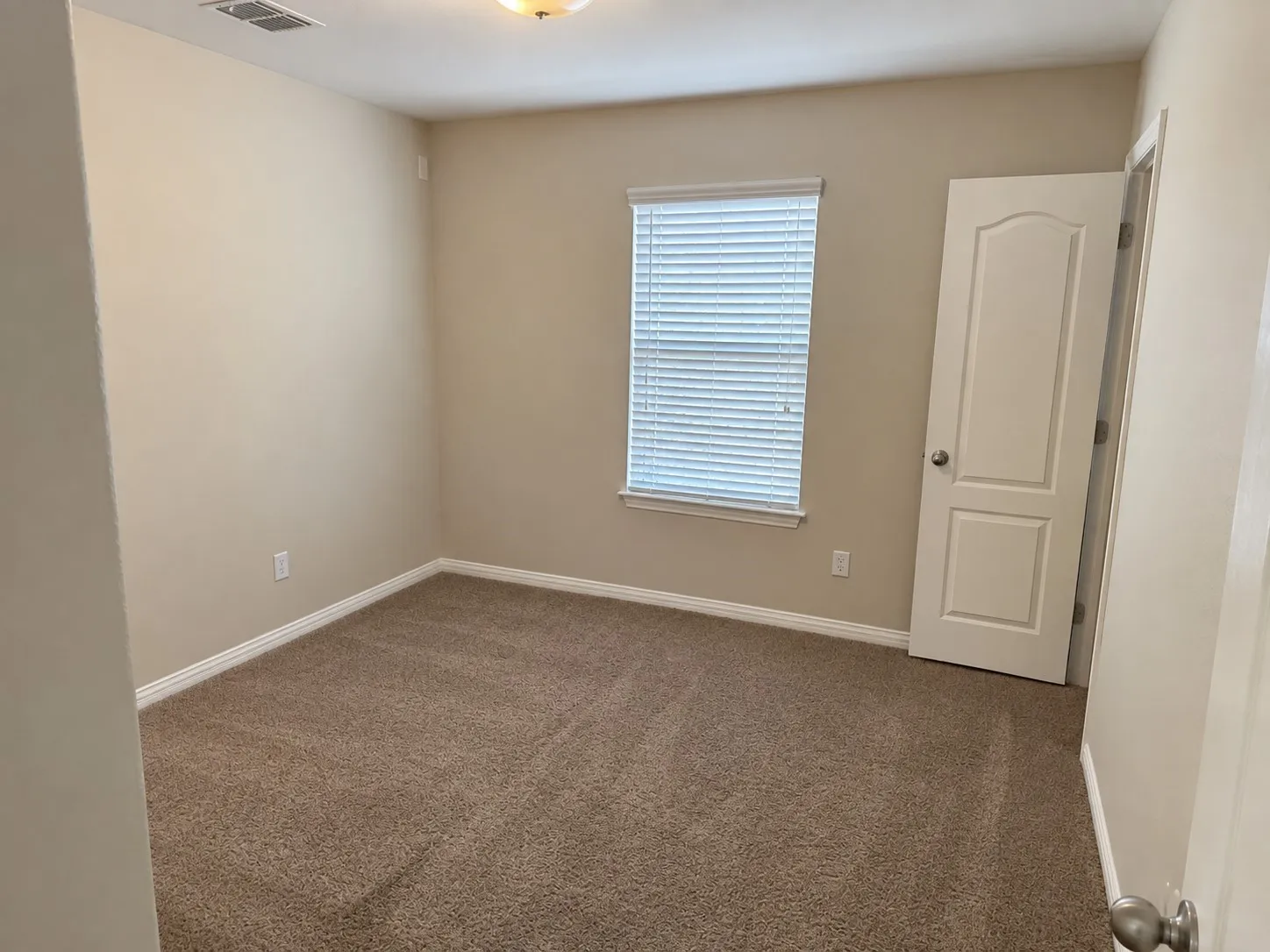 Carpeted flooring, neutral wall tones, window with blinds, overhead lighting, and a white paneled door
