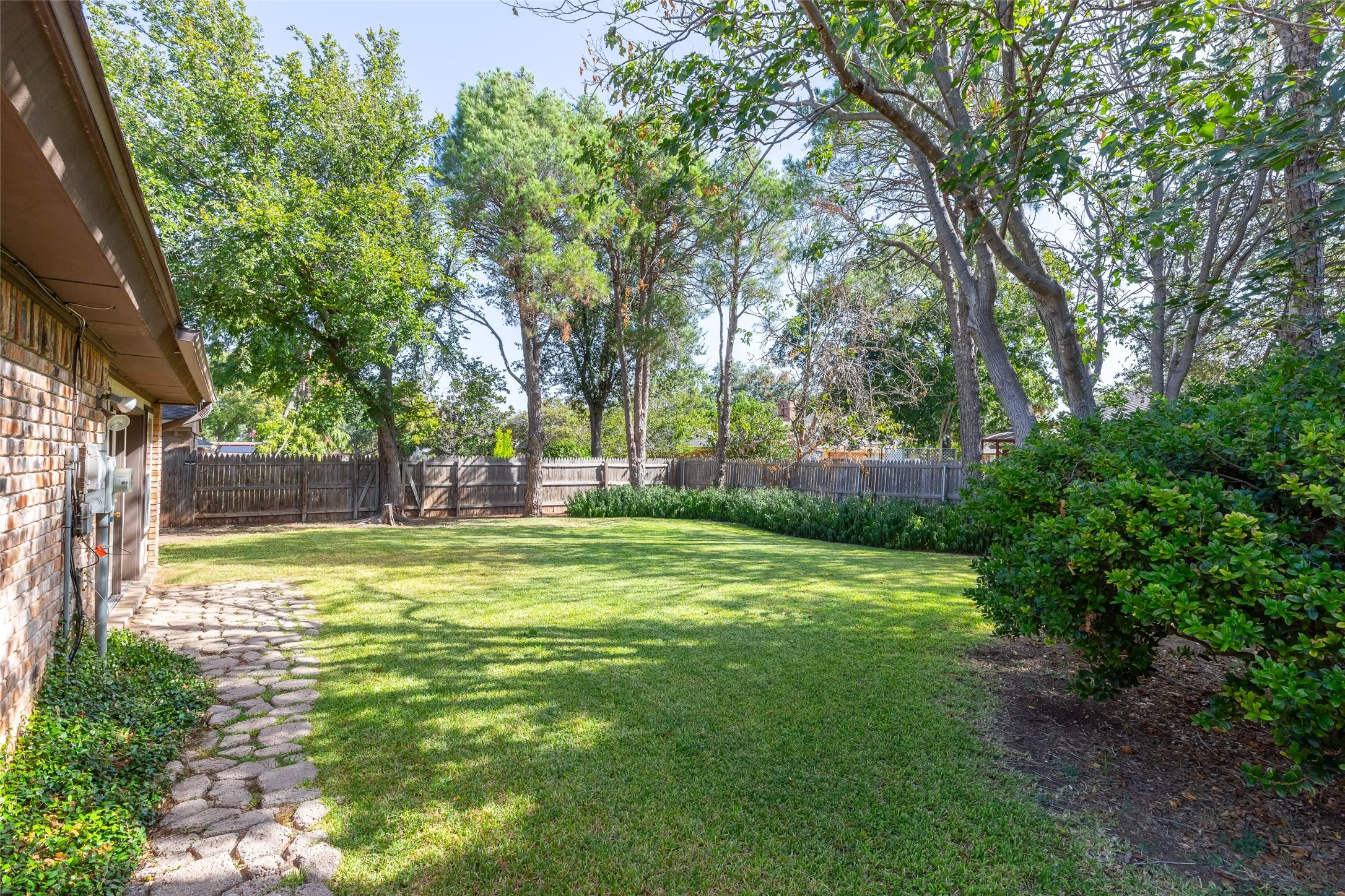 Spacious backyard with a vibrant green lawn, mature trees, wood privacy fencing, and a stone-paved pathway