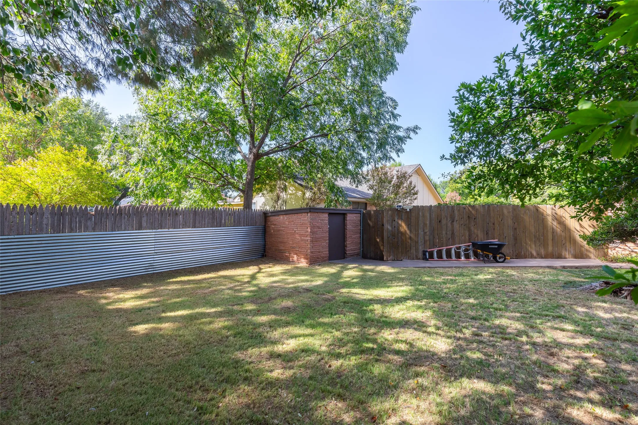 Expansive grassy lawn with a brick exterior storage shed, corrugated metal fencing, and wood plank fencing