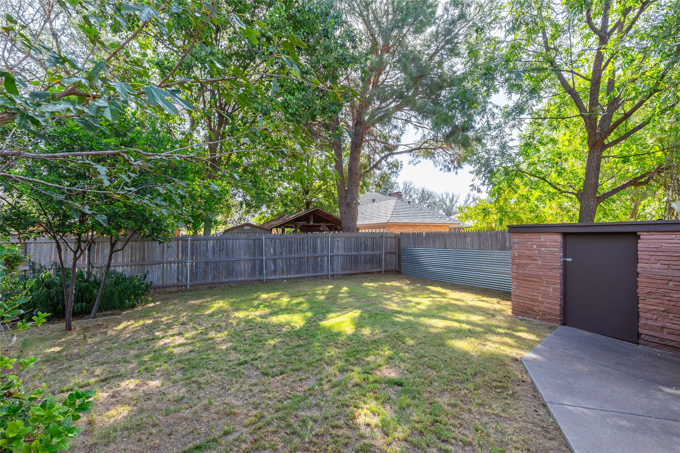 Expansive backyard with a sloped lawn, mature trees, wood fence, and a storage structure featuring a brick facade