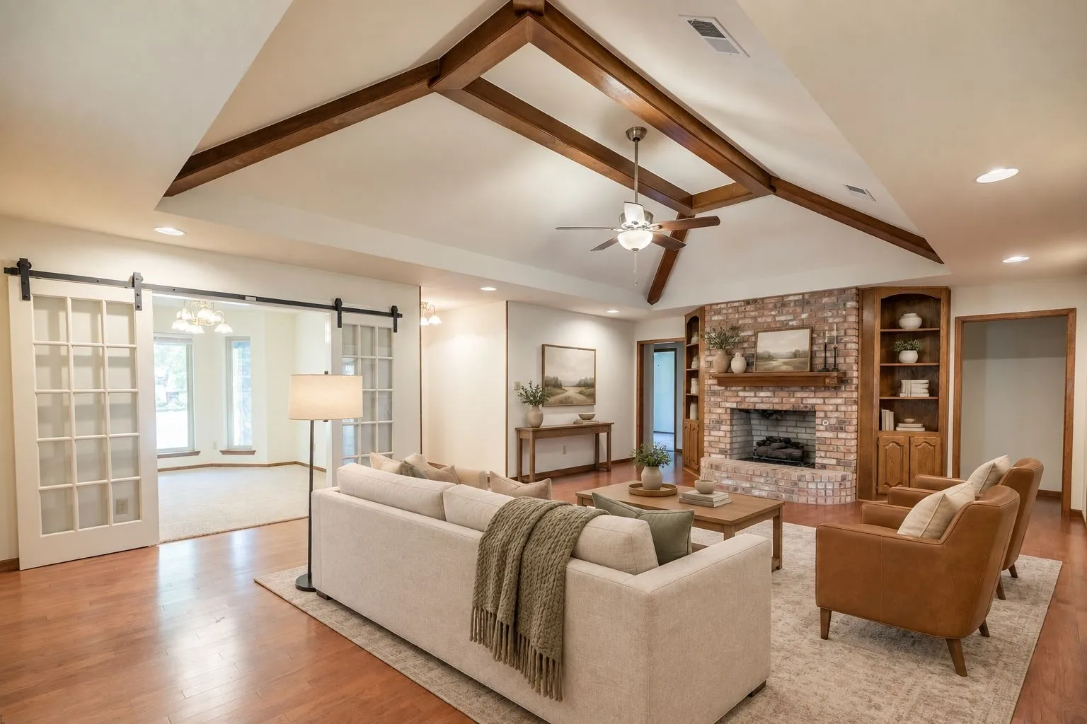 Vaulted ceiling with exposed wood beams, brick-clad fireplace with wood mantel, built-in shelving, wood-finish flooring, and barn door entry