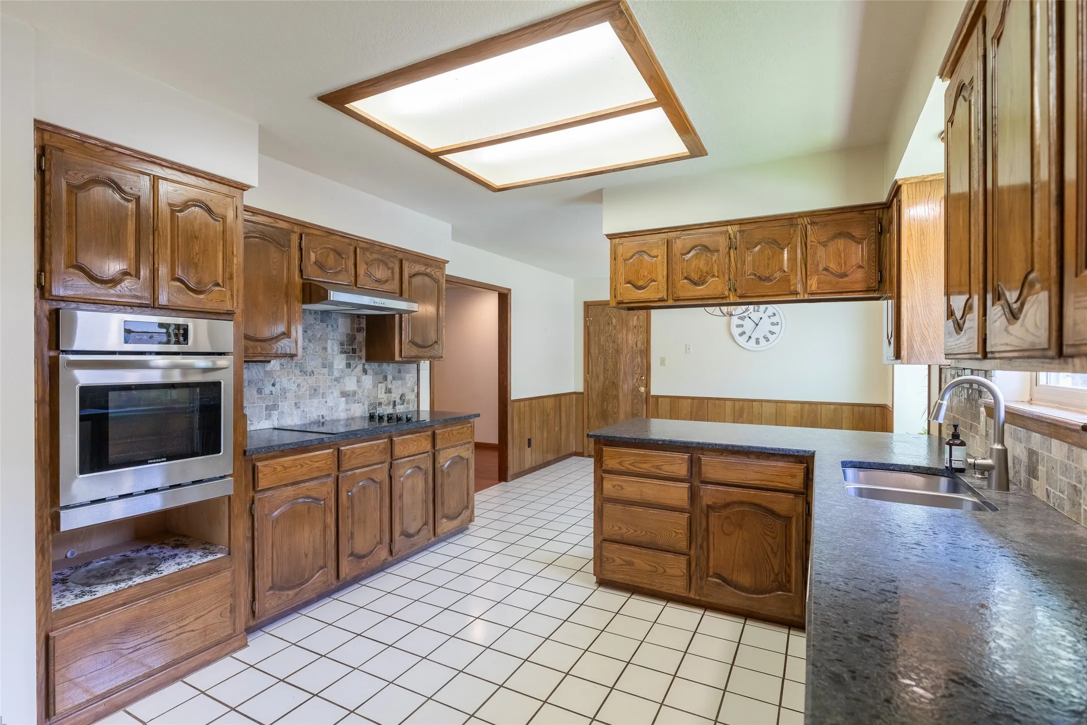 Kitchen featuring wood cabinetry, stainless steel built-in oven, dark countertops, a tiled backsplash, and white square floor tiles