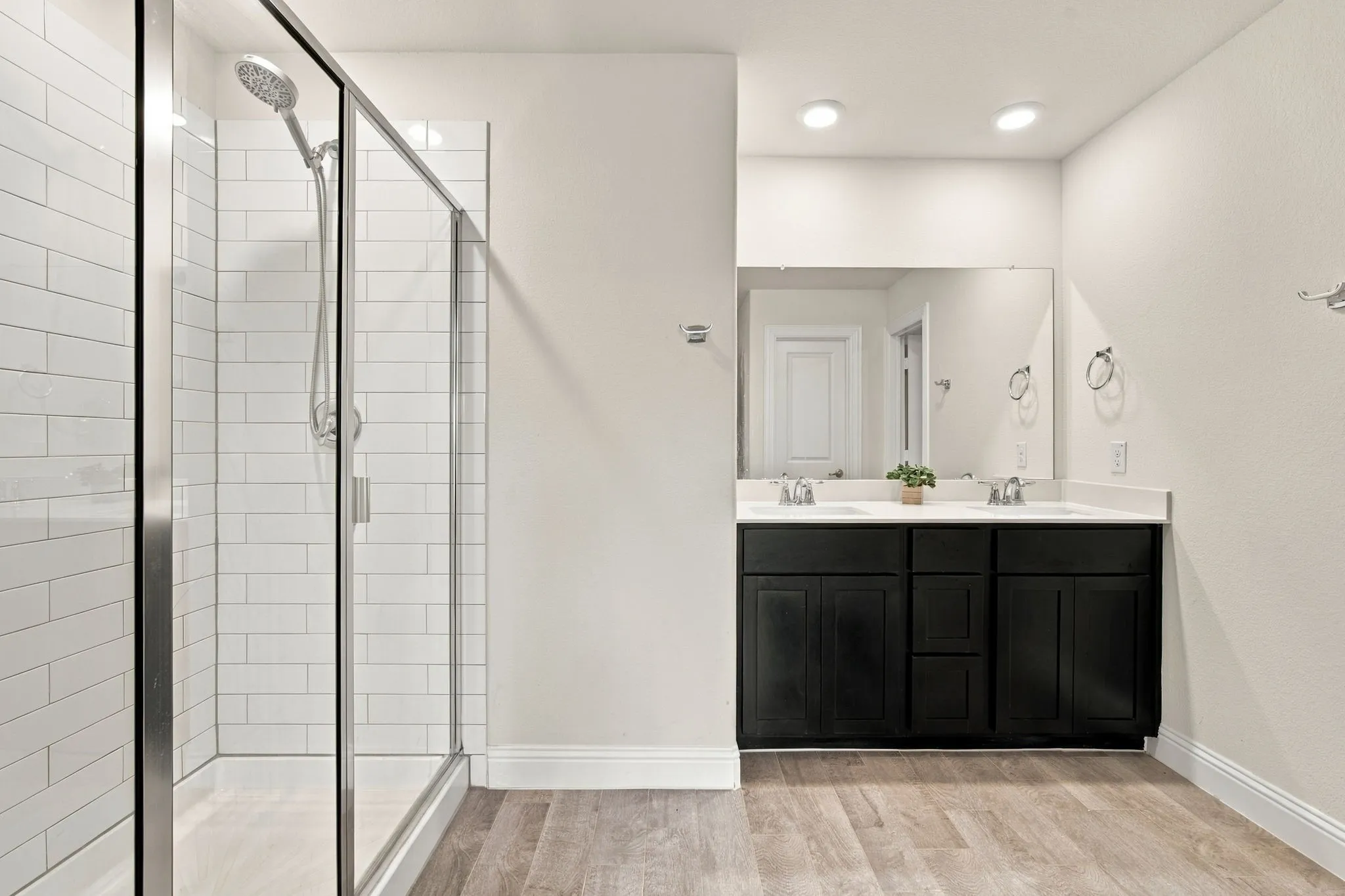 Glass-enclosed shower with subway tile surround, dual vanity with undermount sinks, and wood-finish flooring