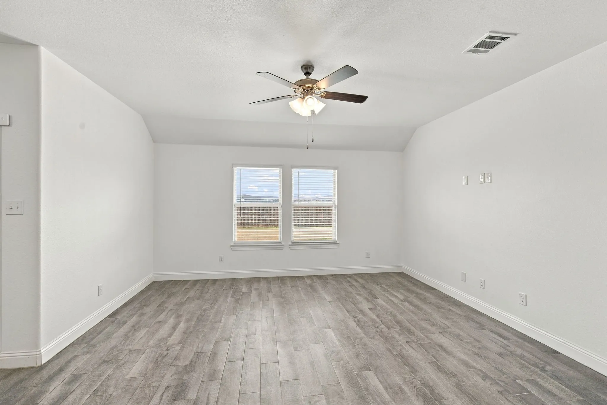 Spacious room featuring wood-finish flooring, two windows with blinds, a ceiling fan with integrated lighting, and white walls