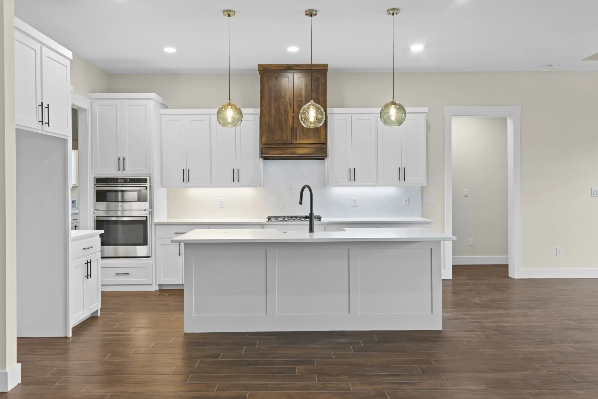 Kitchen featuring white shaker cabinetry, stainless steel double wall ovens, a center island with sink, wood-finish flooring, and pendant lighting