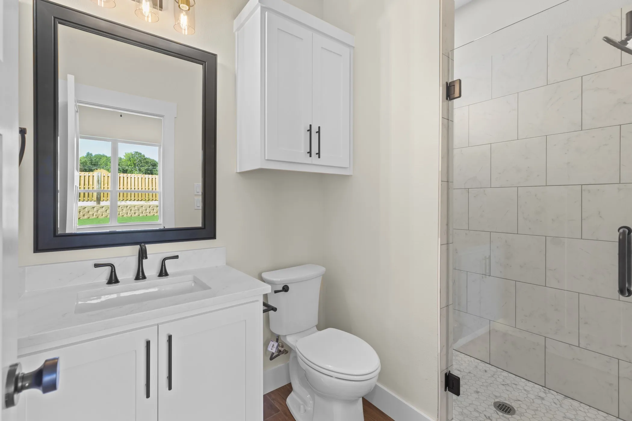 White vanity with an integrated sink and black faucet, framed mirror, built-in storage cabinet, white toilet, and a glass-enclosed shower with tiled walls and a mosaic floor