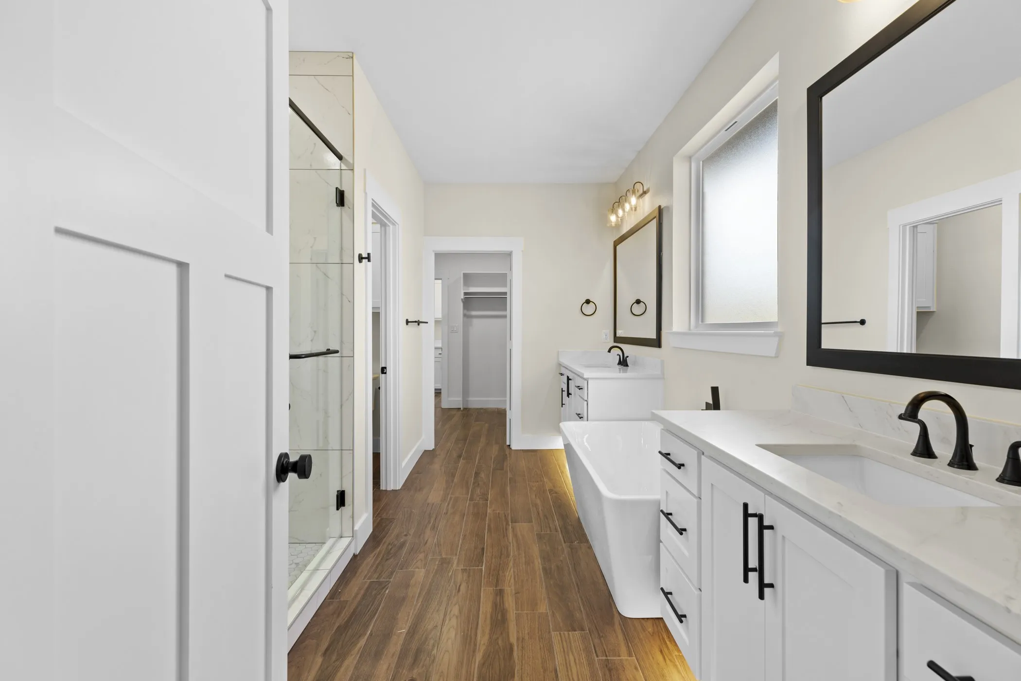 Dual vanity with white cabinetry and matte black hardware, wood-finish flooring, rectangular freestanding soaking tub, glass-enclosed shower, and framed mirrors