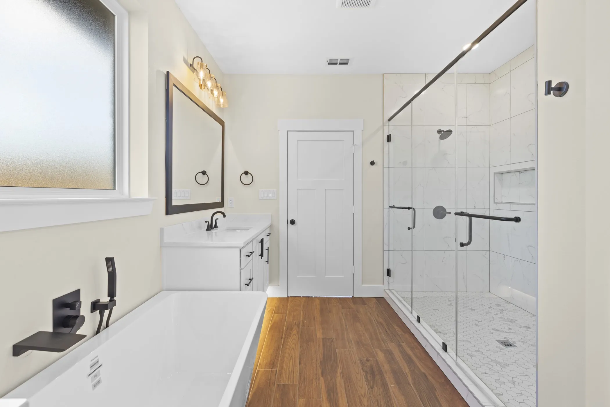 Spacious bathroom featuring a freestanding soaking tub with wall-mounted fixtures, a glass-enclosed shower with tile surround, a single vanity with a white countertop, and wood-finish flooring