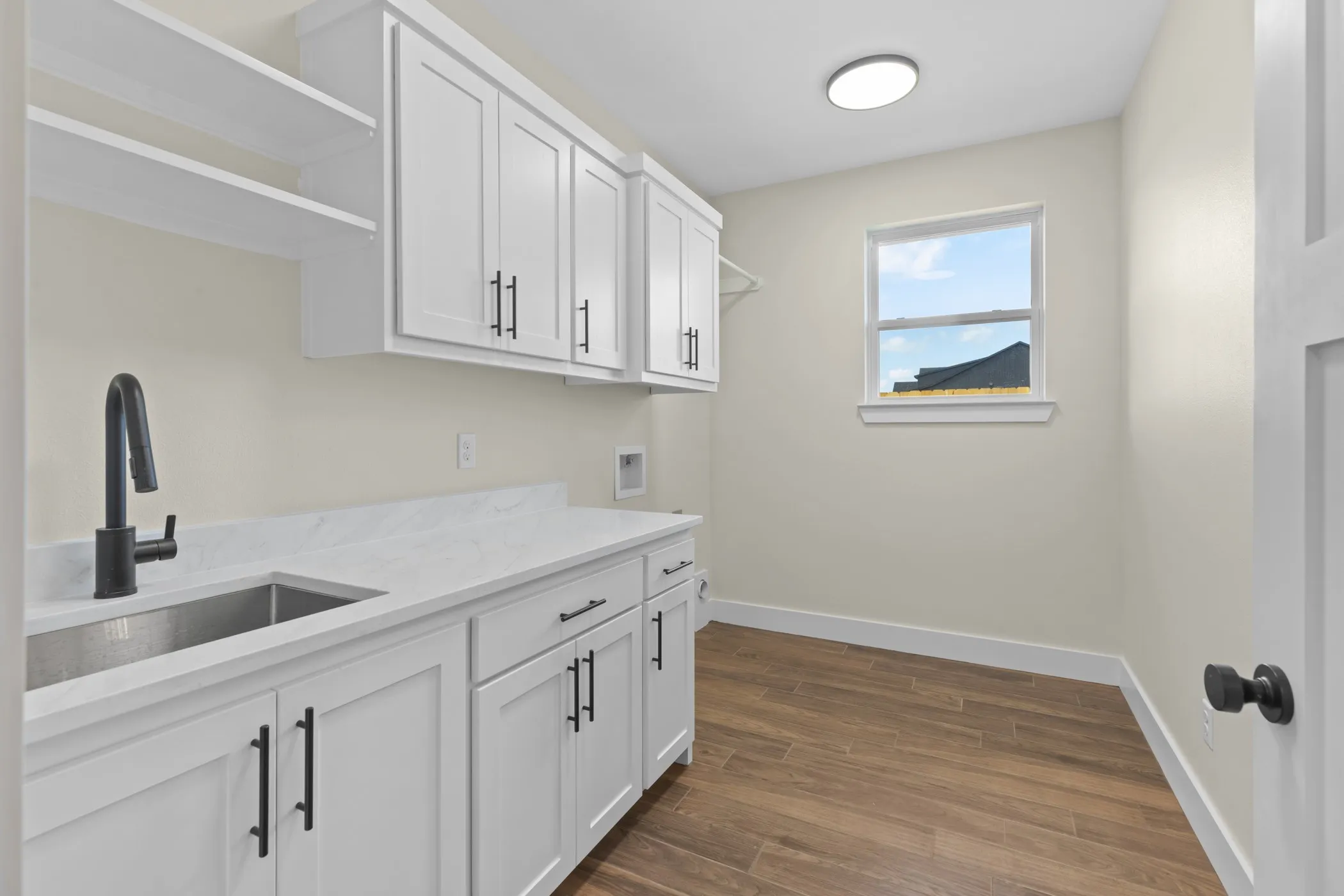 White cabinetry with matte black hardware, integrated stainless steel sink, expansive white countertop, wood-finish flooring, and a single window