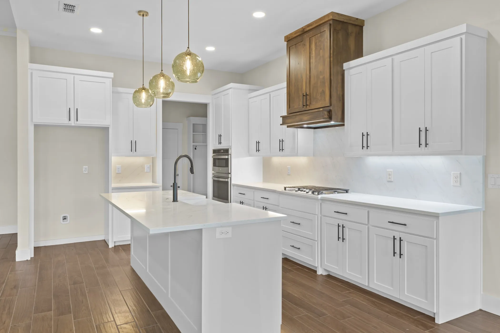 Kitchen featuring a center island with a sink, white cabinetry with dark hardware, a wood-finish range hood, double wall ovens, and wood-finish flooring