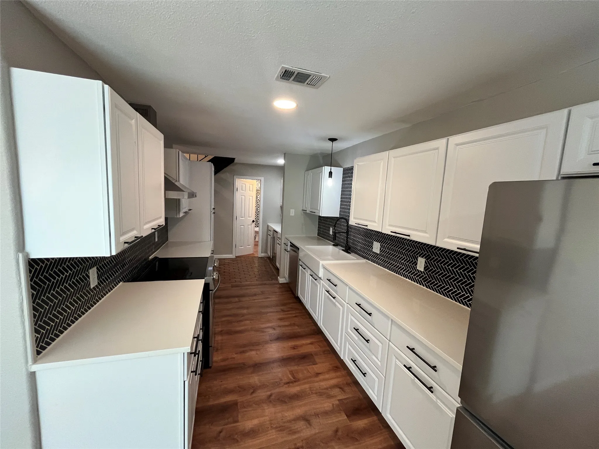Galley kitchen featuring white cabinetry, quartz-finish countertops, herringbone tile backsplash, wood-finish flooring, and a stainless steel appliance suite