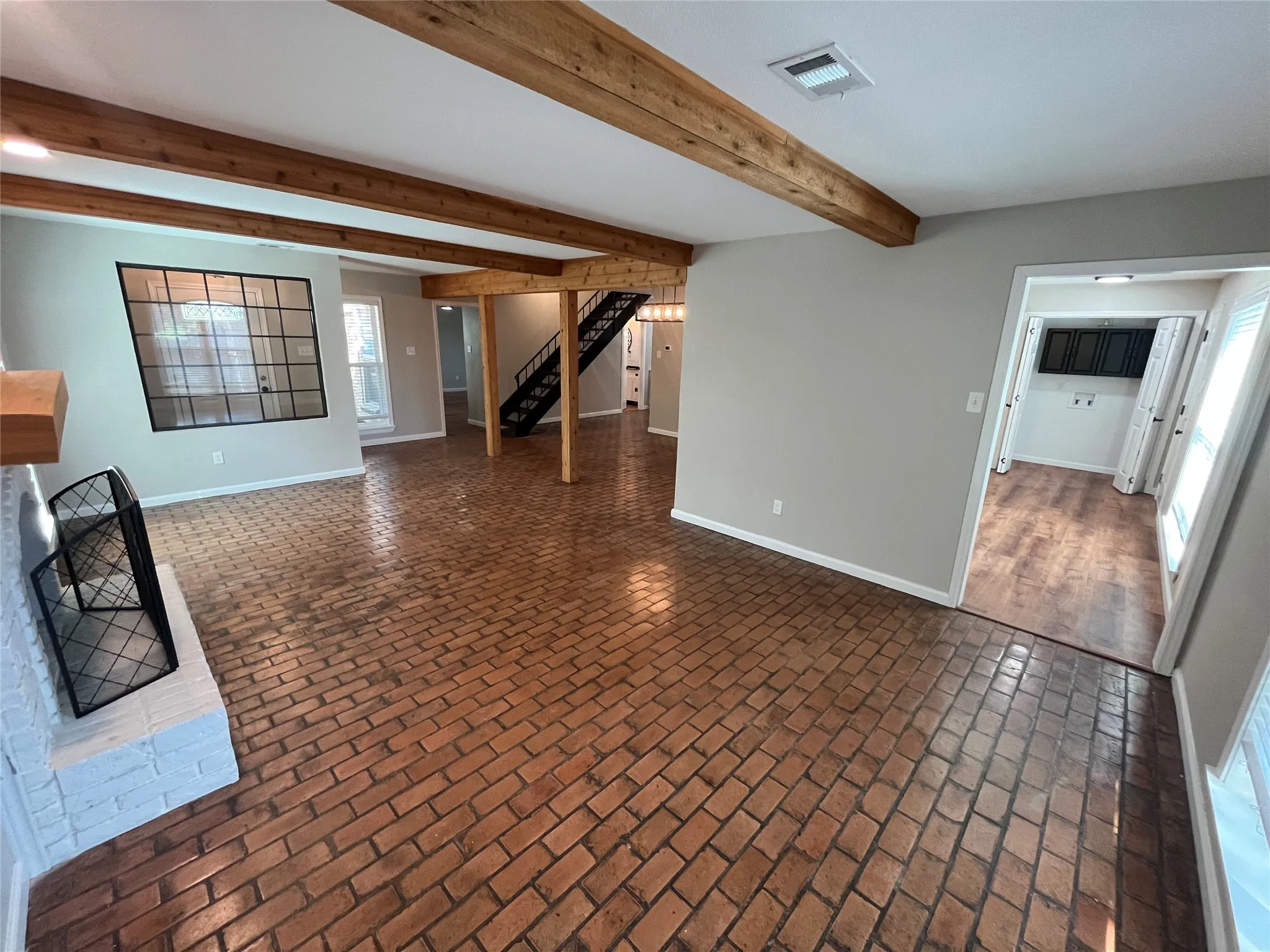 Brick flooring, exposed wood beams, white brick fireplace, built-in window with black grid, and wood-finish flooring in an adjoining room