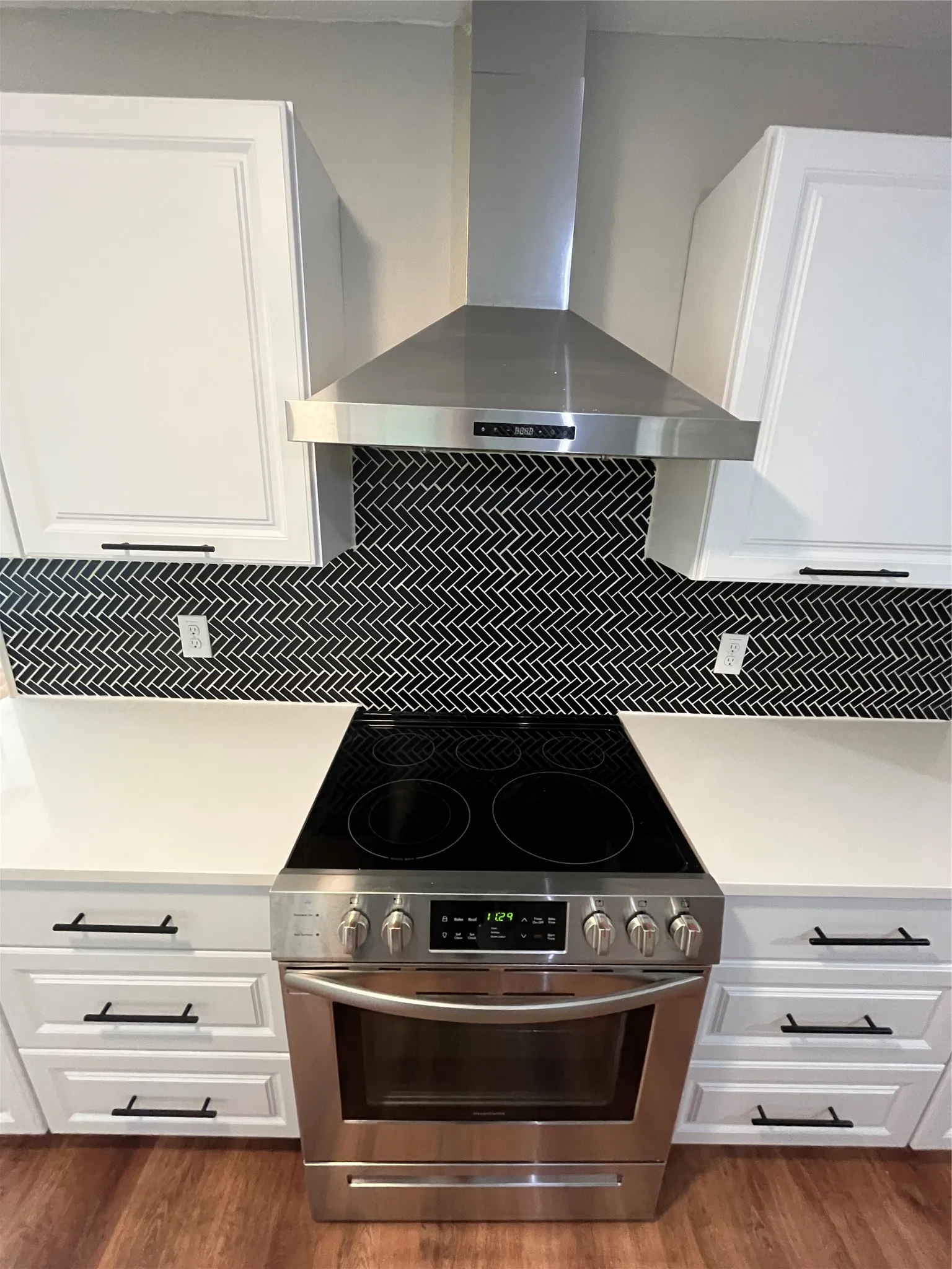 Stainless steel range with a smooth cooktop, white shaker-style cabinetry, black herringbone tile backsplash, white countertops, and black hardware