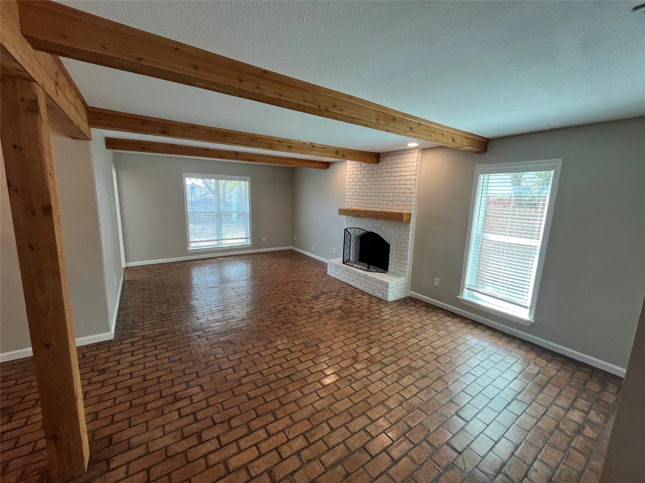 Exposed wood beams, wood-finish flooring, white brick fireplace with a wood mantel, and multiple windows