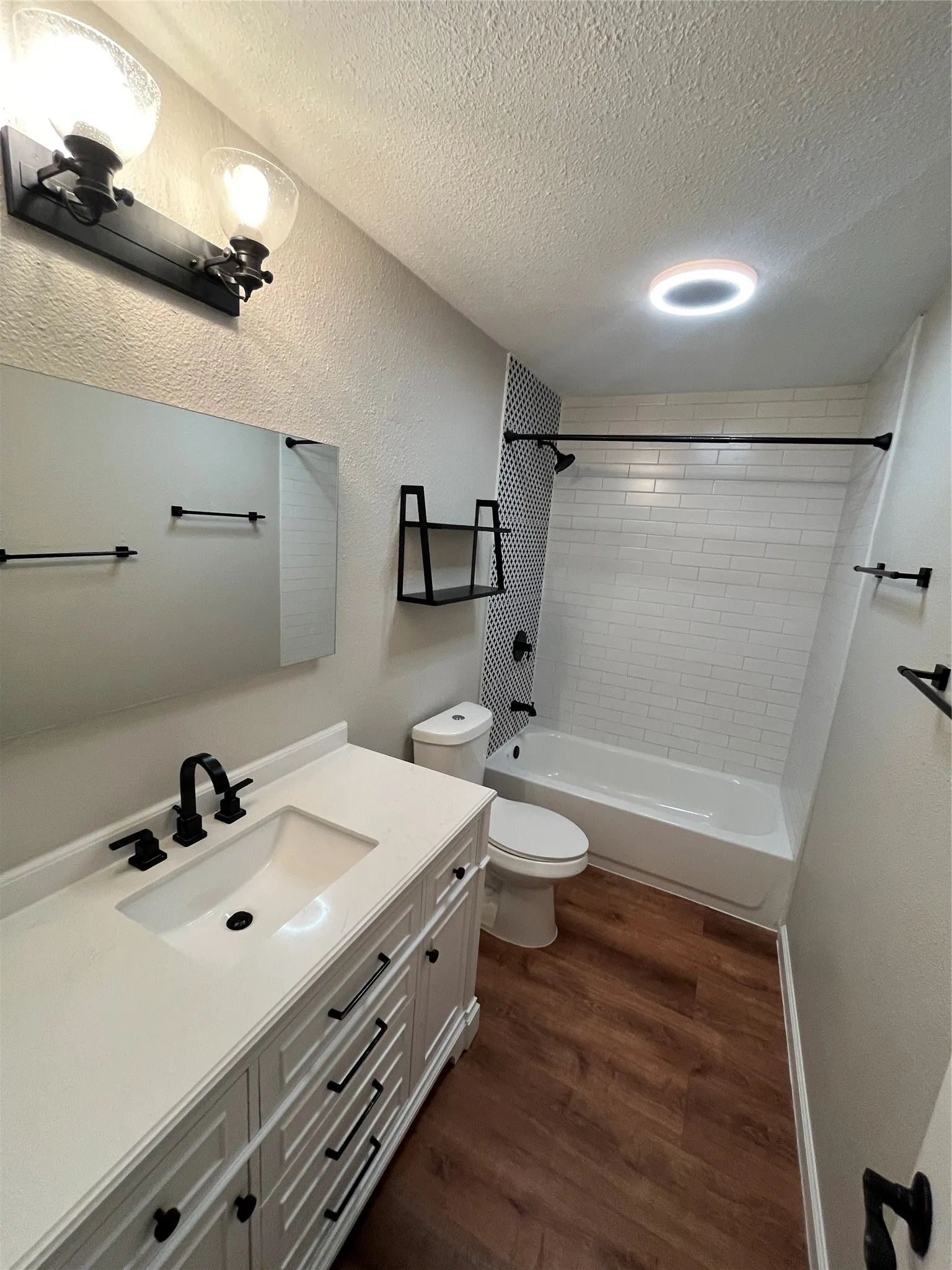 White vanity with a rectangular sink and matte black hardware, wood-finish flooring, subway tile bath surround, and a coordinating black shower rod