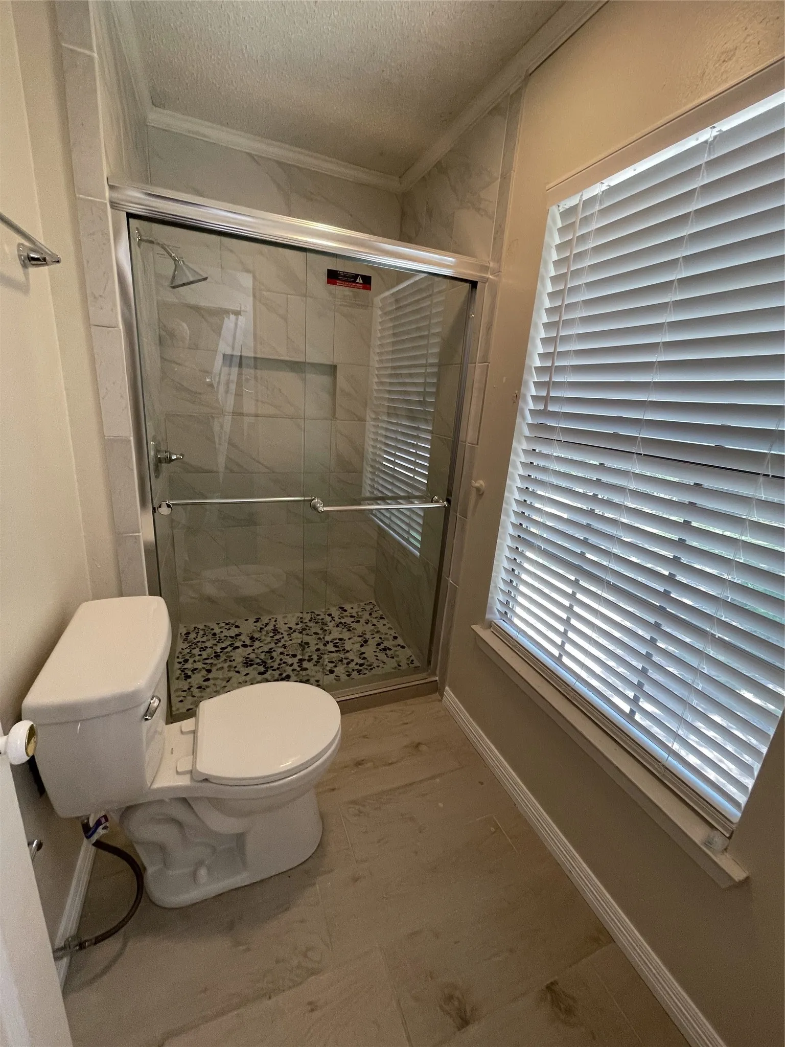 Glass-enclosed shower with tiled walls and pebble flooring, wood-finish flooring, and a window with blinds