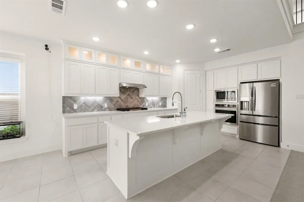White cabinetry with glass-front uppers, stainless steel appliances, a center island with sink, herringbone tile backsplash, and recessed lighting