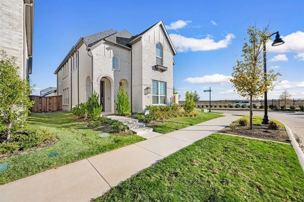 White brick exterior with arched entry, dark shingle roof, mature landscaping, and concrete walkways