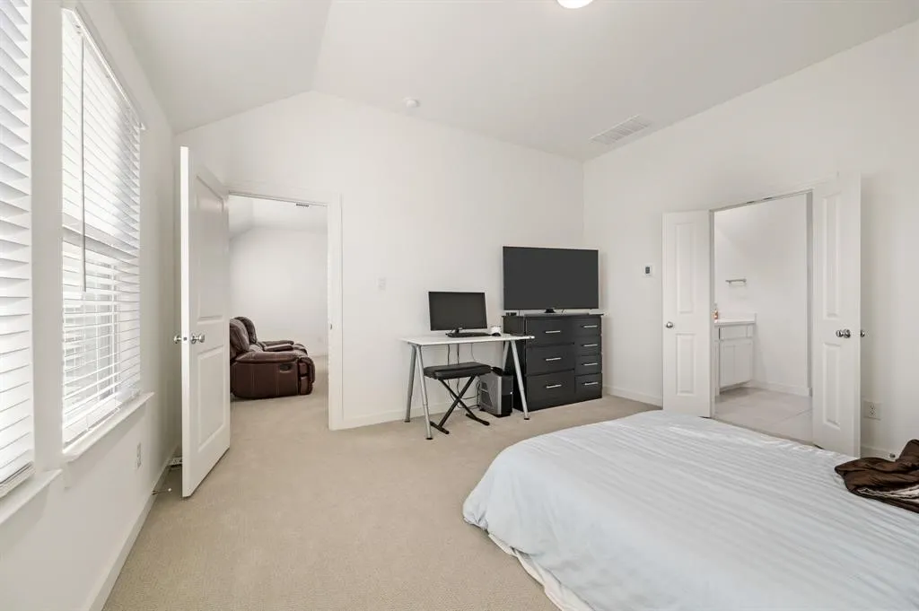 Vaulted ceiling with recessed lighting, carpet flooring, dual windows with blinds, and double doors opening to a bathroom with a vanity