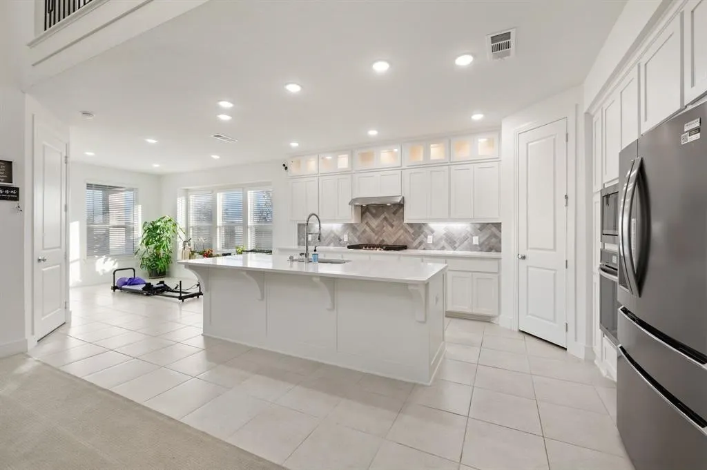 Modern kitchen featuring a large island with an undermount sink, stainless steel appliances, white cabinetry with display uppers, a herringbone tile backsplash, and recessed lighting