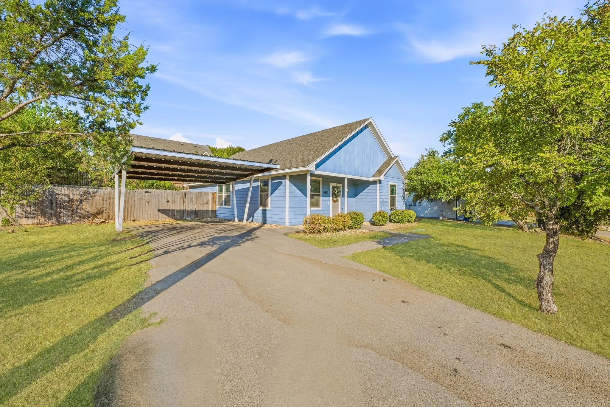 Blue siding exterior with a dark shingle roof, covered carport, gravel driveway, and surrounding green lawn