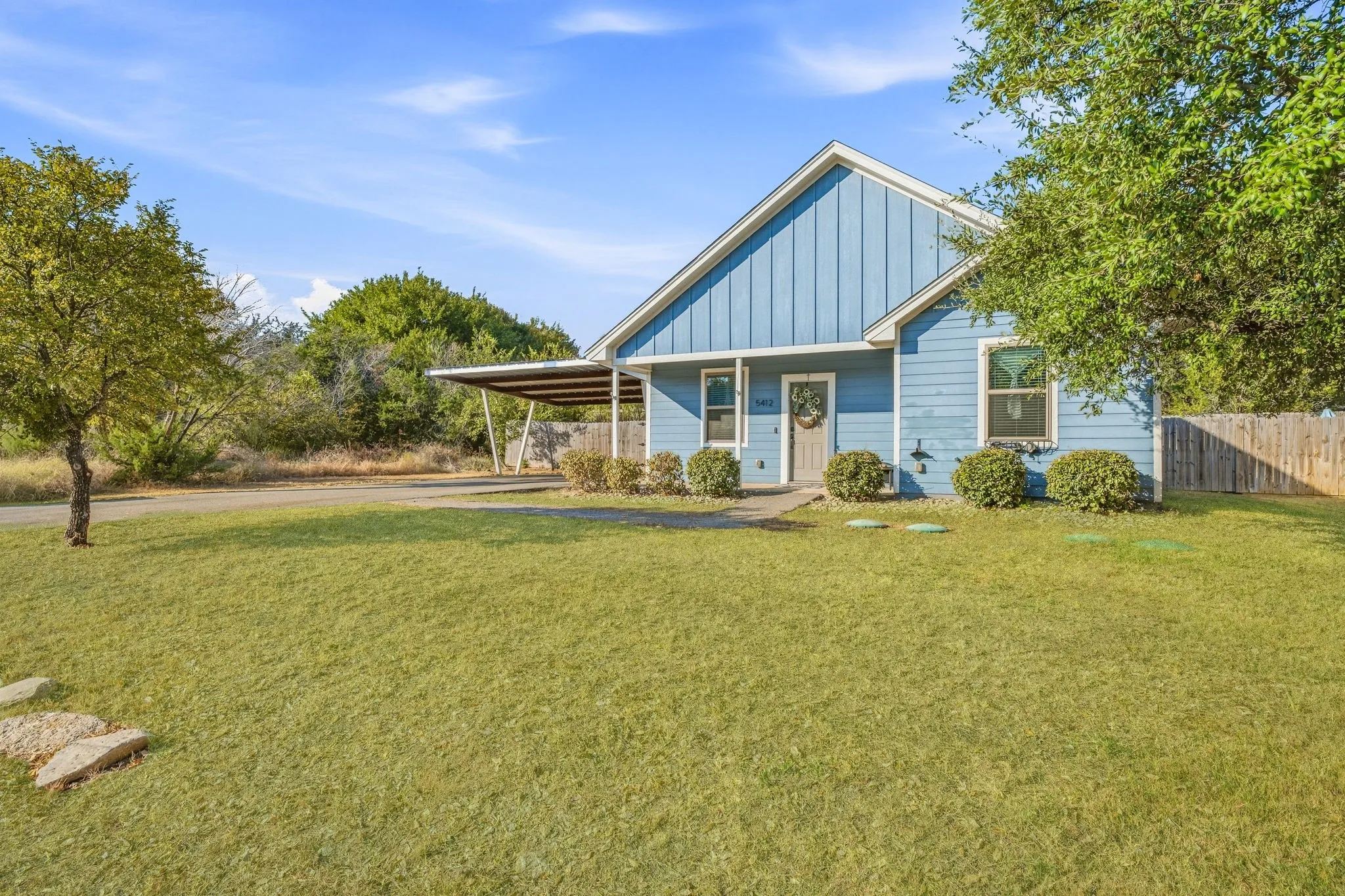 Blue exterior siding, carport structure, front yard with green lawn, and mature trees