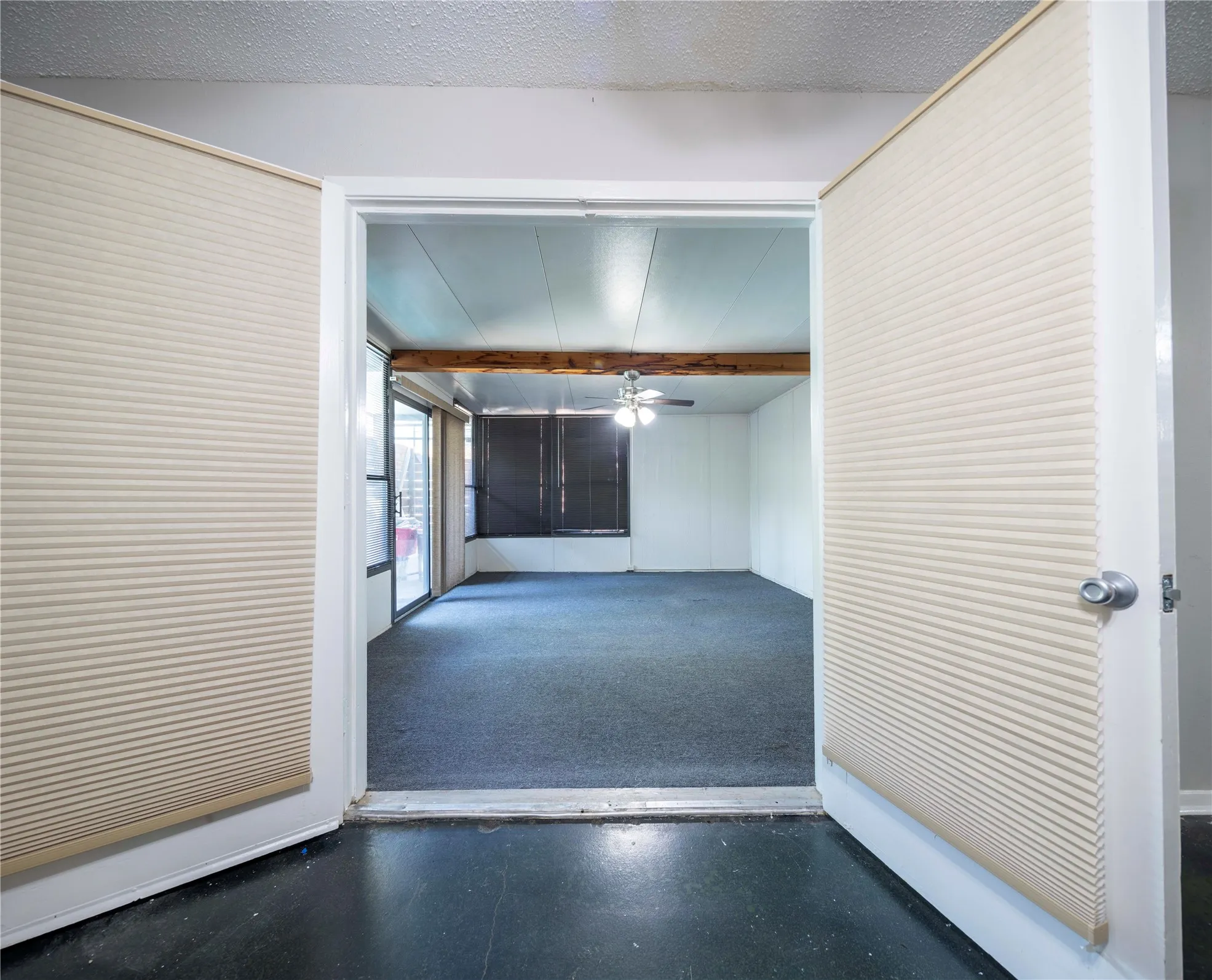 Room with dark carpeting, exposed wood beam, ceiling fan with light fixture, and sliding glass doors with dark screens