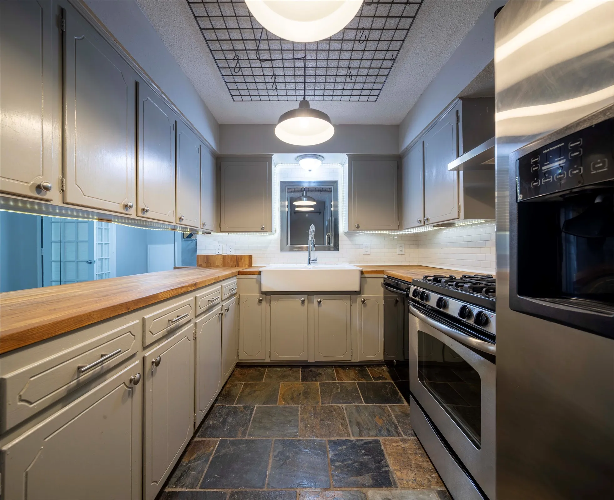 Kitchen featuring slate-style flooring, light-colored cabinetry with modern hardware, wood-finish countertops, a white farmhouse-style sink, and stainless steel appliances