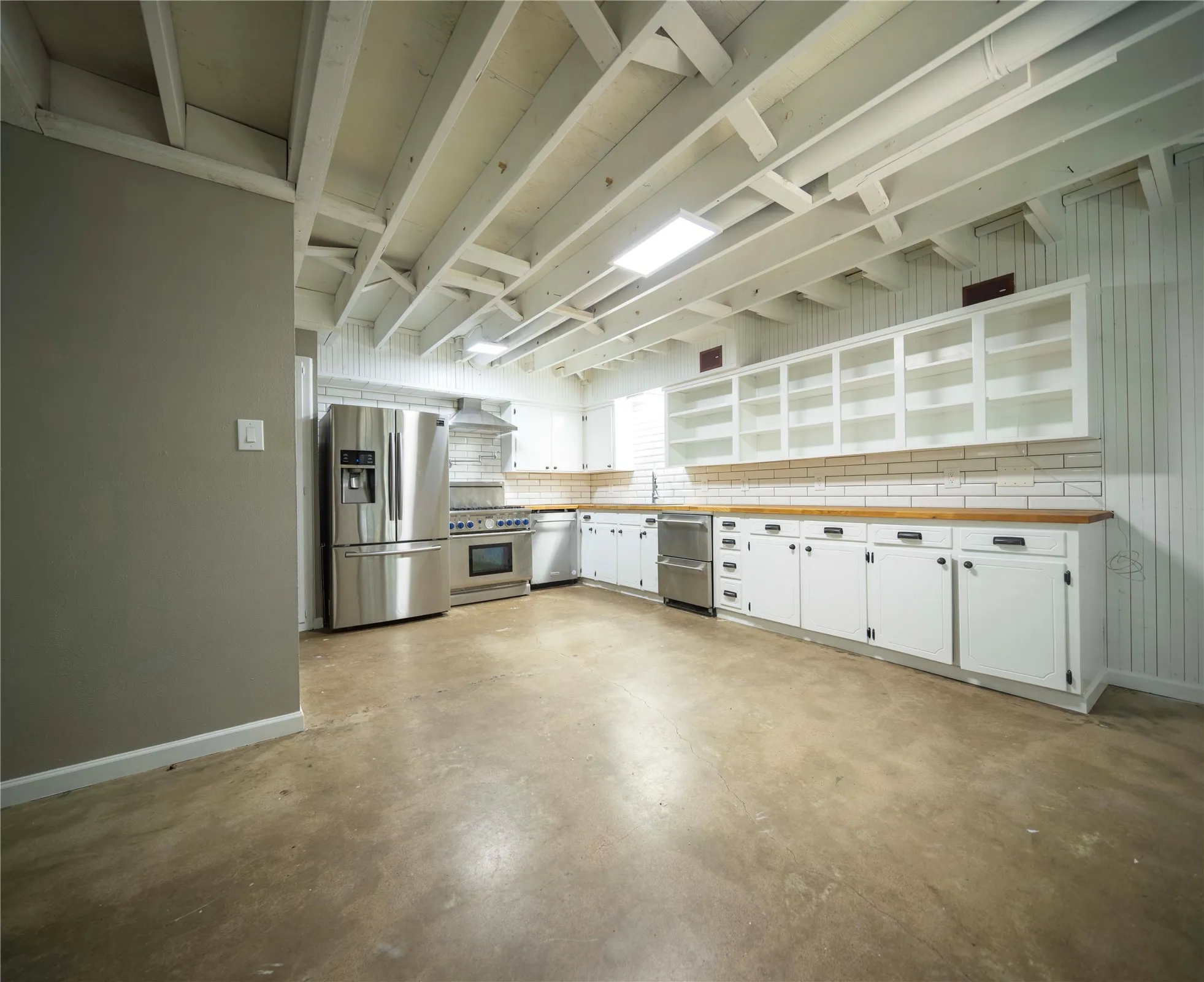 Exposed ceiling joists with recessed lighting, stainless steel kitchen appliances, white shaker-style cabinetry with dark hardware, wood-finish countertops, and subway tile backsplash