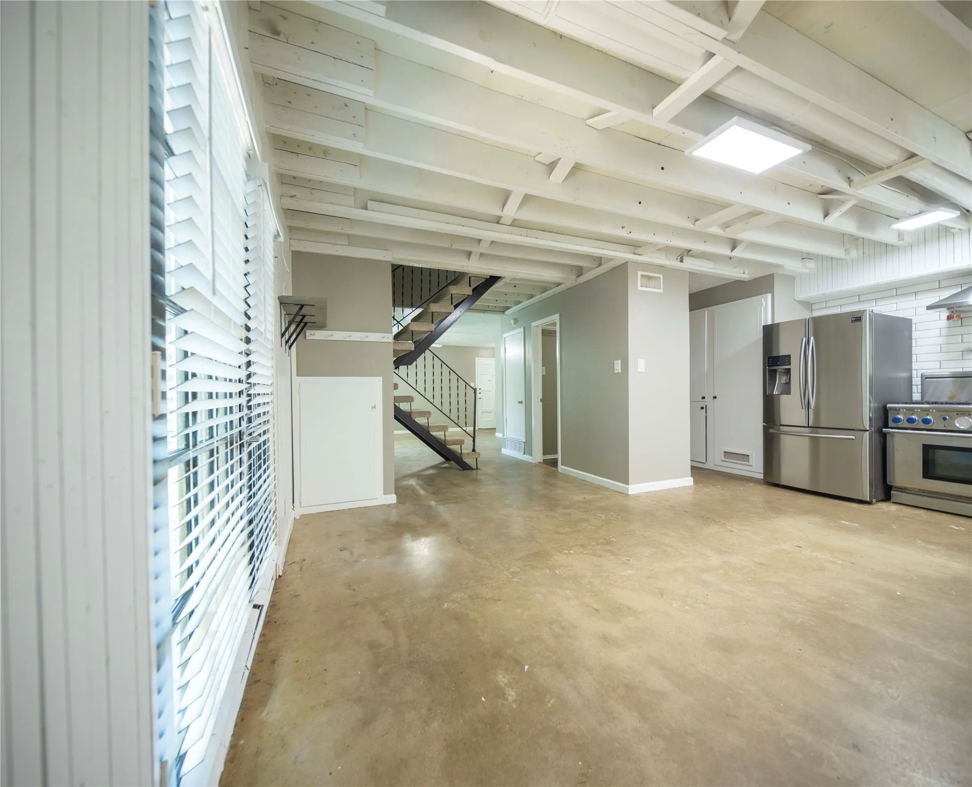 Exposed beam ceiling, polished concrete flooring, stainless steel appliances, white brick backsplash, and a black wrought iron staircase